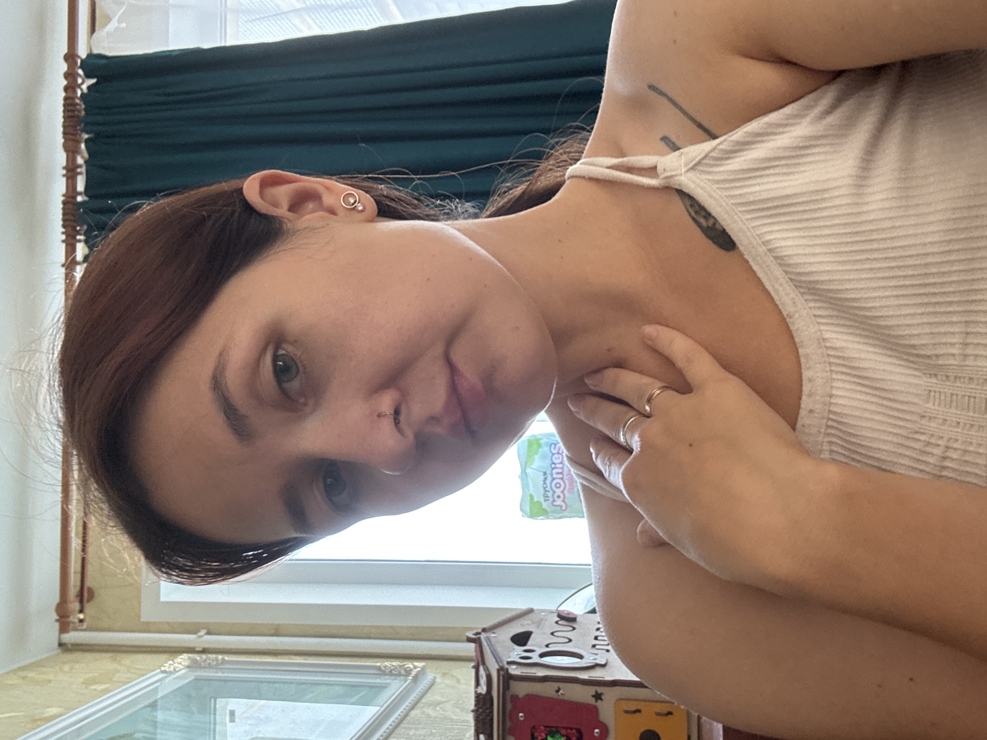 A woman with auburn hair pulled back in a ponytail and green eyes is taking a selfie indoors. She is wearing a white ribbed tank top and has a nose ring and an earring in her left ear. Her right hand is resting on her neck, and she is wearing two rings on her right hand. There is a tattoo on her left shoulder. In the background, there is a wooden toy, a package of baby wipes, and a green curtain. The image appears to have been taken in an apartment in Naberezhnye Chelny, Russia, given the context provided.