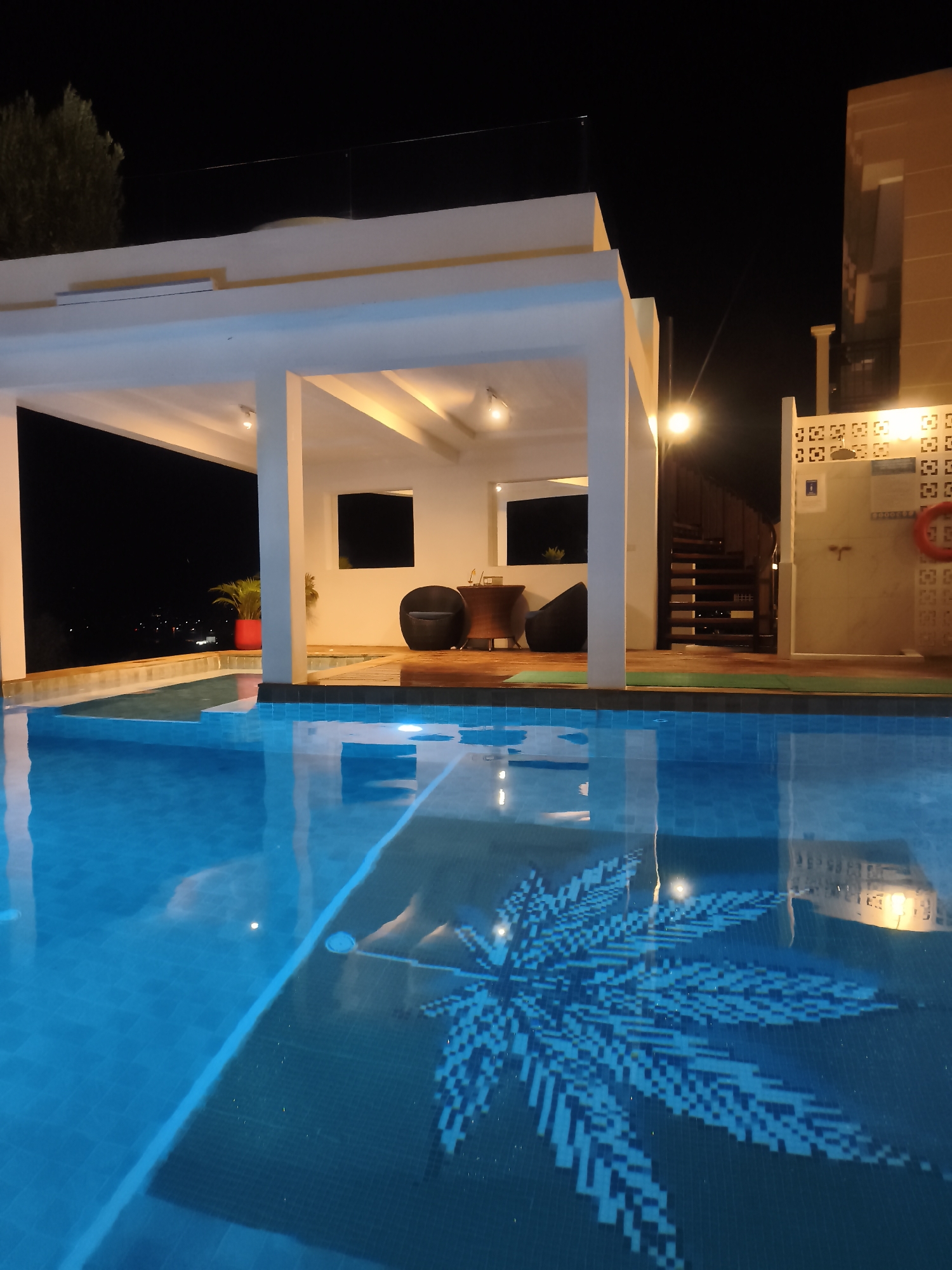 This is a night-time, eye-level, outdoor shot of a swimming pool and a covered patio area. The pool is illuminated and reflects the lights from the patio and surrounding structures. A mosaic of a leaf pattern is visible on the bottom of the pool. To the left of the pool is a white, open-sided structure with a flat roof, supported by white pillars, with lights illuminating the ceiling. Inside this structure, a dark chair and beanbag chairs are visible, along with a wooden table. Beyond the patio, a spiral staircase leads upwards, and a wall with a decorative pattern and an orange life preserver is visible. The background is dark, indicating night, with some distant lights visible on a hillside. The image does not provide information about its specific location in Baan Bang Rak, Thailand.