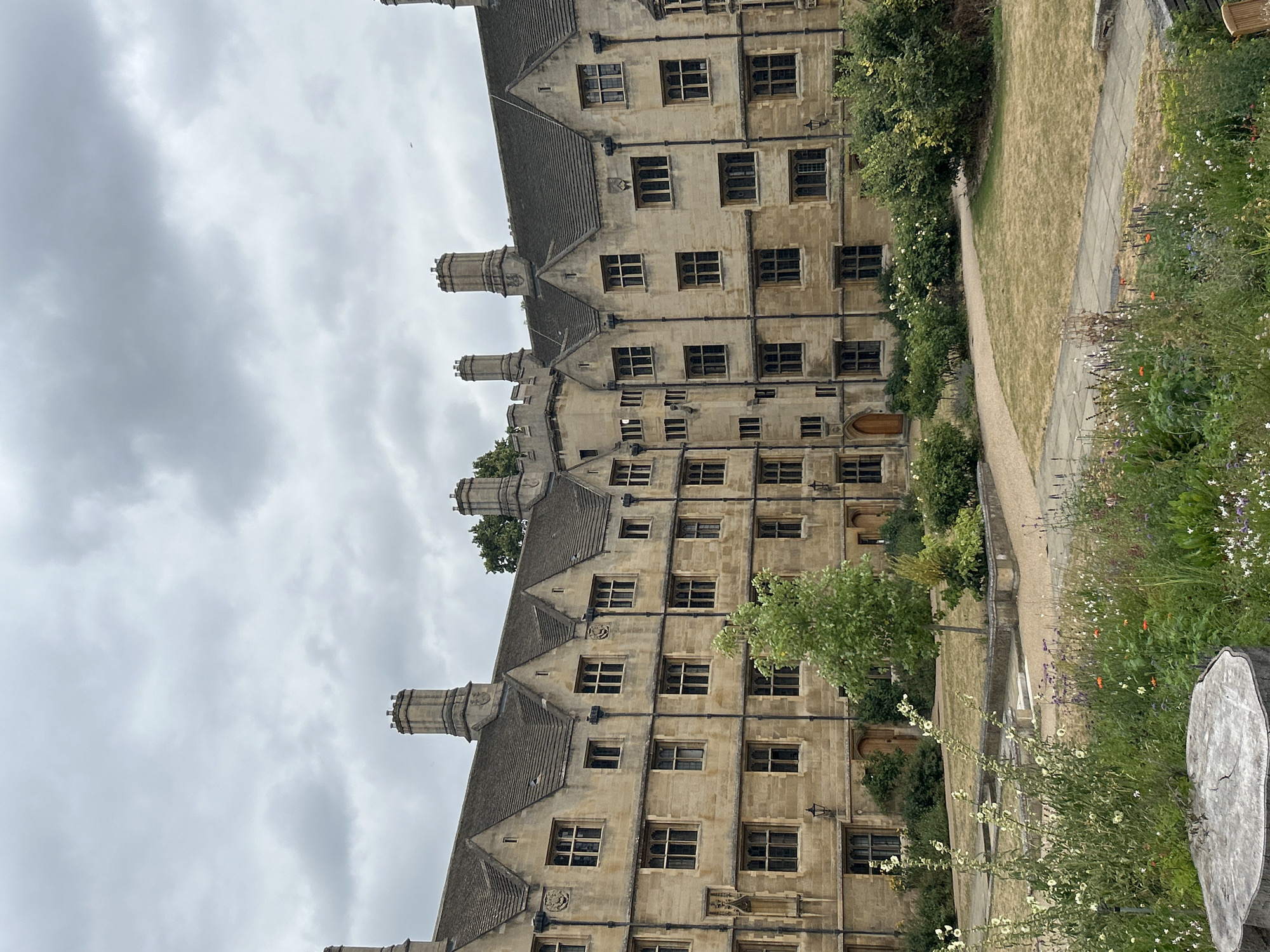 The image depicts a view of a quadrangle at St John's College, Cambridge, United Kingdom. The scene is dominated by a large, multi-story, stone building with a steeply pitched, tiled roof and numerous windows. The architecture is characterized by its classical style, with ornate chimneys and decorative stonework. In the foreground, a garden area with a variety of flowers and a tree is visible. The ground is covered in patchy grass and paved walkways. The sky is overcast with grey clouds. There are no people or animals visible in the image.