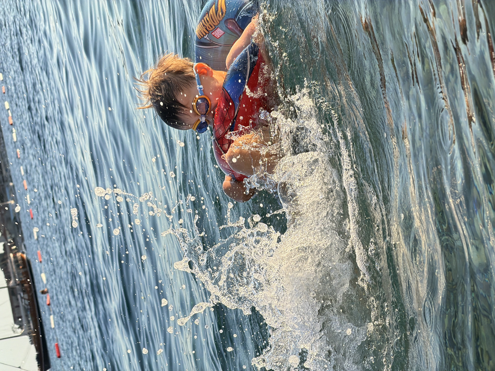 A young child wearing swimming goggles and a Spiderman-themed inflatable vest is in the sea, splashing water. The child is holding onto a red and blue inflatable toy, possibly a ring or a float. The water is blue and appears somewhat choppy, with sunlight reflecting off the surface, creating a shimmering effect. Large water splashes are visible in the foreground, indicating active movement. In the background, out of focus, are several boats anchored in the water and a line of buoys. The overall scene suggests a recreational activity at the beach or a harbor in Budva, Montenegro, during daylight.
