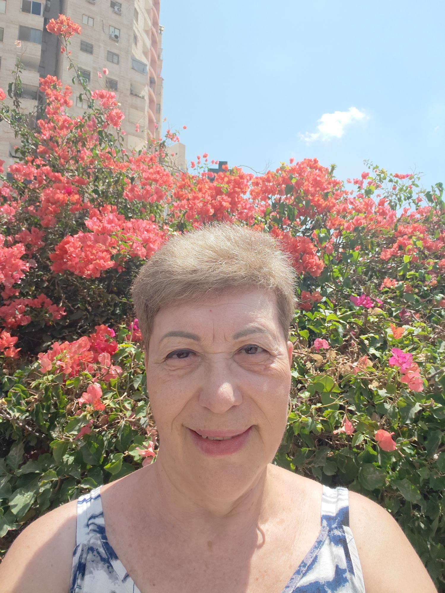 A middle-aged woman with short brown hair and a slight smile takes a selfie in front of a building and a large bush of vibrant pink and red bougainvillea flowers. She is wearing a white top with a blue abstract pattern. The background includes a light-colored multi-story building with visible windows and balconies, and a bright blue sky with a single white cloud. The scene appears to be outdoors, likely in Be'er Sheva, Israel, given the distinctive building architecture and flora commonly found in the region.