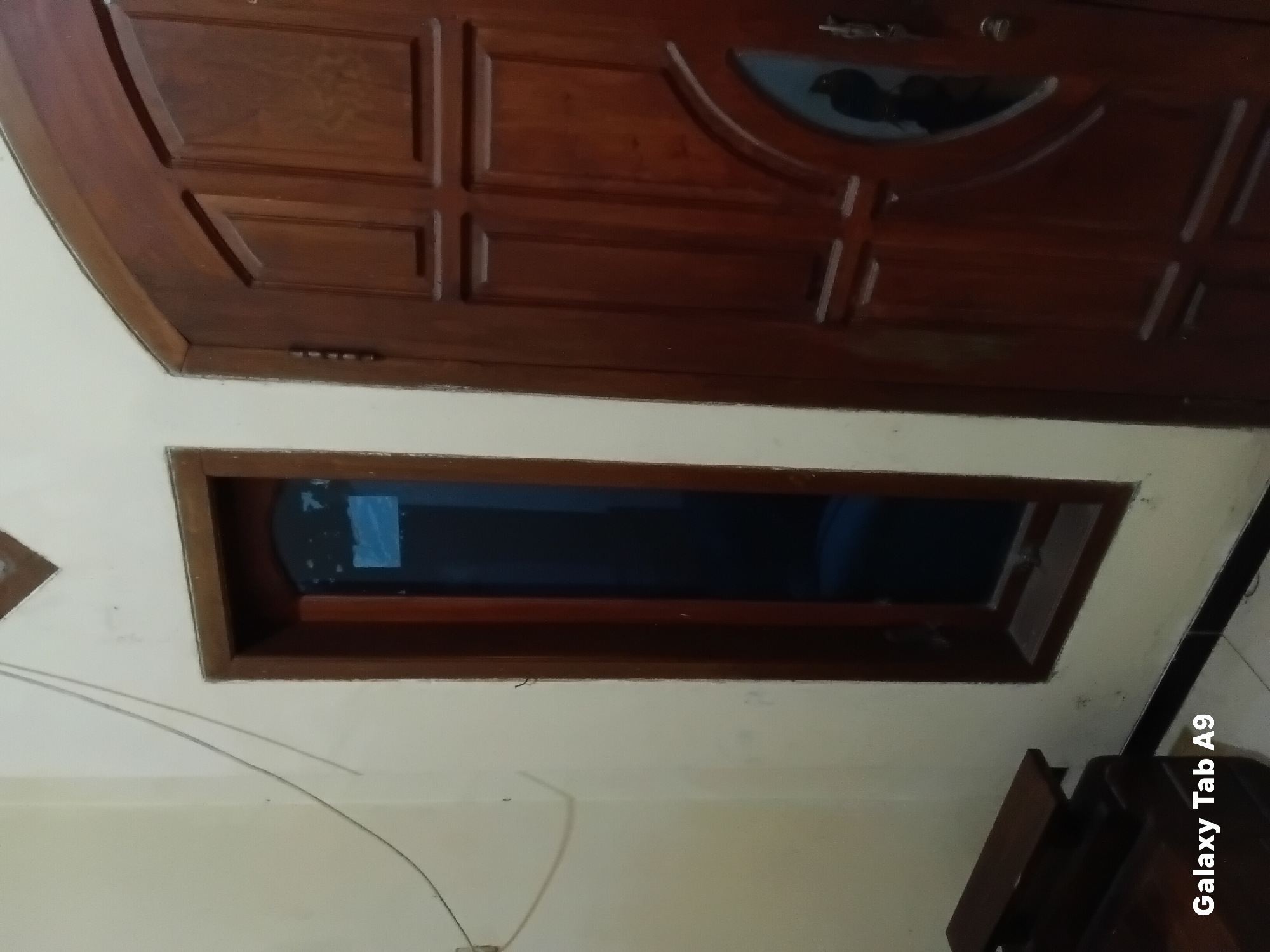 The image shows a close-up view of a wooden door and a narrow window in a room. The door is made of dark brown wood with carved panels and a decorative oval glass insert. The window is tall and narrow with a wooden frame, and the glass appears to be tinted dark blue. A portion of a wooden bench is visible in the foreground on the left side of the image. A thin wire runs diagonally across the wall above the window and door. The wall is painted a light cream color. The floor is tiled with dark and light tiles. The bottom left corner of the image displays the text "Galaxy Tab A9". The scene appears to be indoors.
