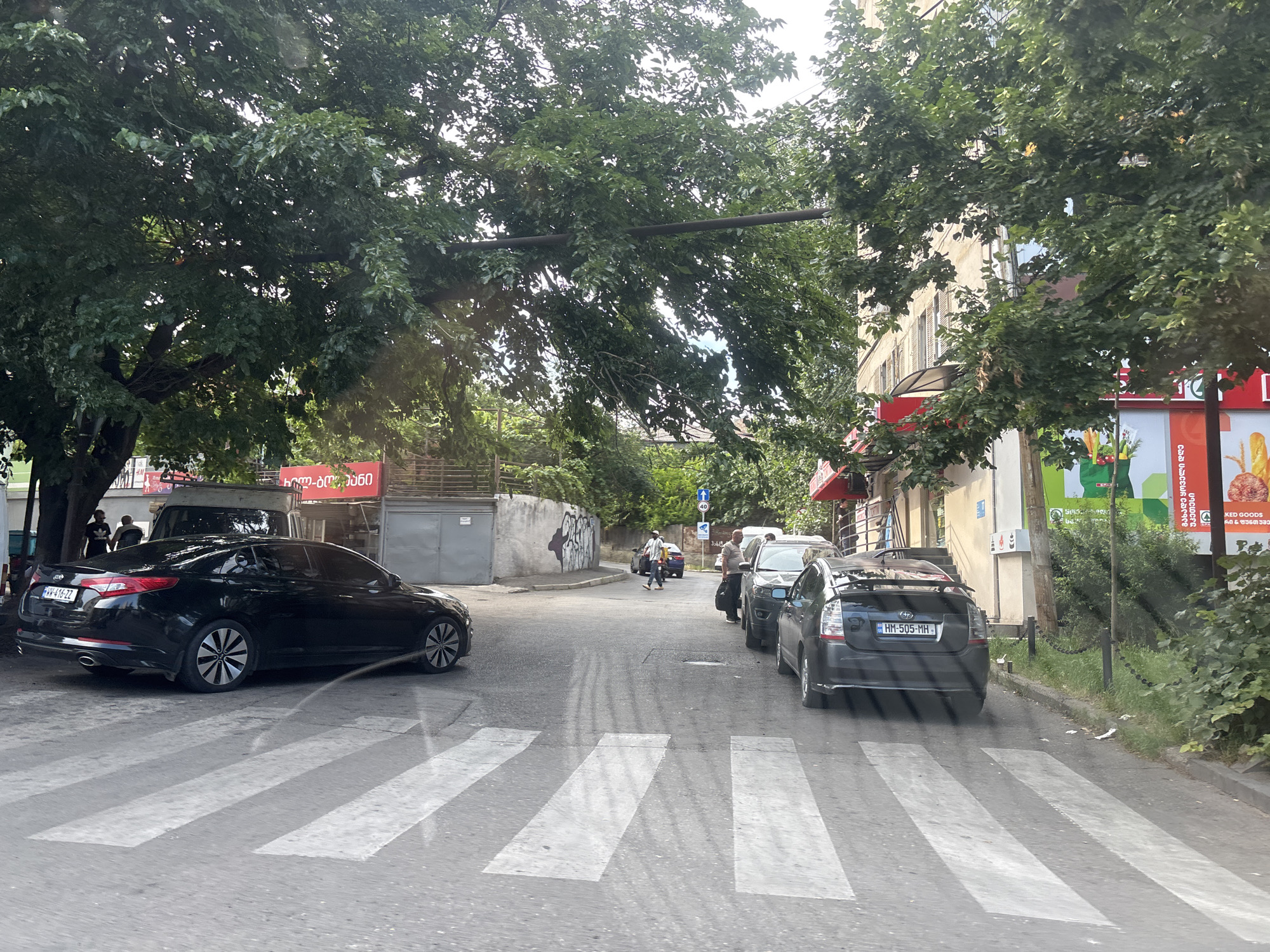 This media file captures a street scene in Tbilisi, Georgia. The foreground is dominated by a pedestrian crossing. On the left, a black Kia Optima is parked, partially obscuring the crossing. Beyond the Kia, a large tree with dense green foliage extends across the upper portion of the image, casting shadows and dappled light.
Further down the street, several cars are visible, including a dark-colored Prius. In the background, buildings line the right side of the street, with some showing commercial signage. A gray wall with graffiti marks the left side of the road. The sky is overcast, suggesting a possibly humid or cloudy day.
Several people are present, mostly walking or standing near the parked vehicles. One person is seen walking across the pedestrian crossing in the middle distance. Another individual appears to be standing next to the black Kia.
Visible text includes license plates on the cars, "WW-416-ZZ" on the Kia and "HM-505-MH" on the Prius. There is also text in Georgian on the building to the right, with snippets like "გახადე ყოველი დღე უკეთესი" (Make every day better) and "KED GOODS". A circular sign with "40" indicates a speed limit. The overall scene depicts a typical urban environment with vehicles, pedestrians, and commercial establishments.