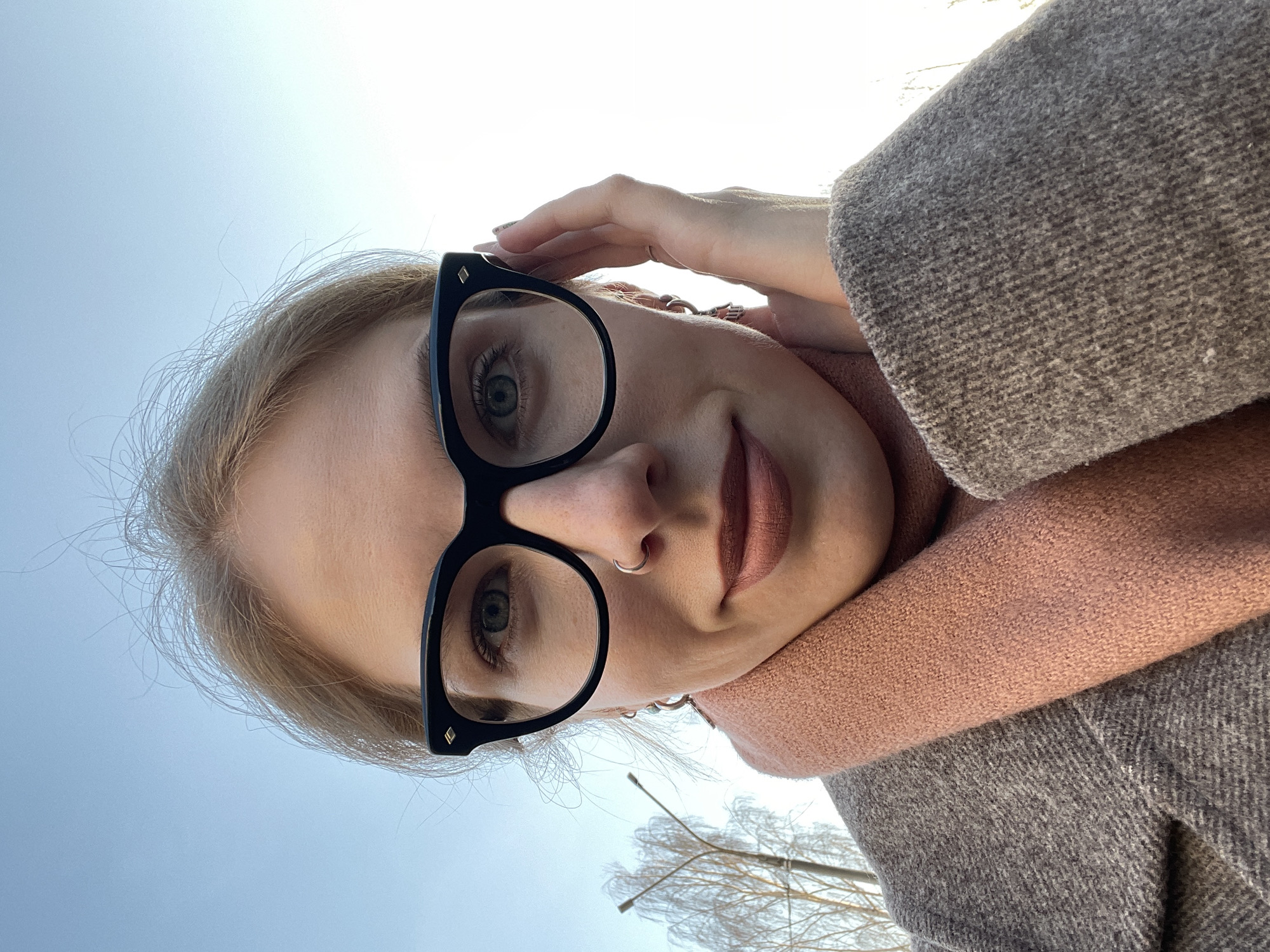 A woman takes a selfie in Izhevsk, Russia. She is wearing large black-framed glasses and a pink scarf, which contrasts against her gray woolen coat. She has a nose ring and is touching her forehead with her right hand. The background shows a pale blue sky and bare trees suggesting winter. The lighting is bright, and the perspective is looking upwards.