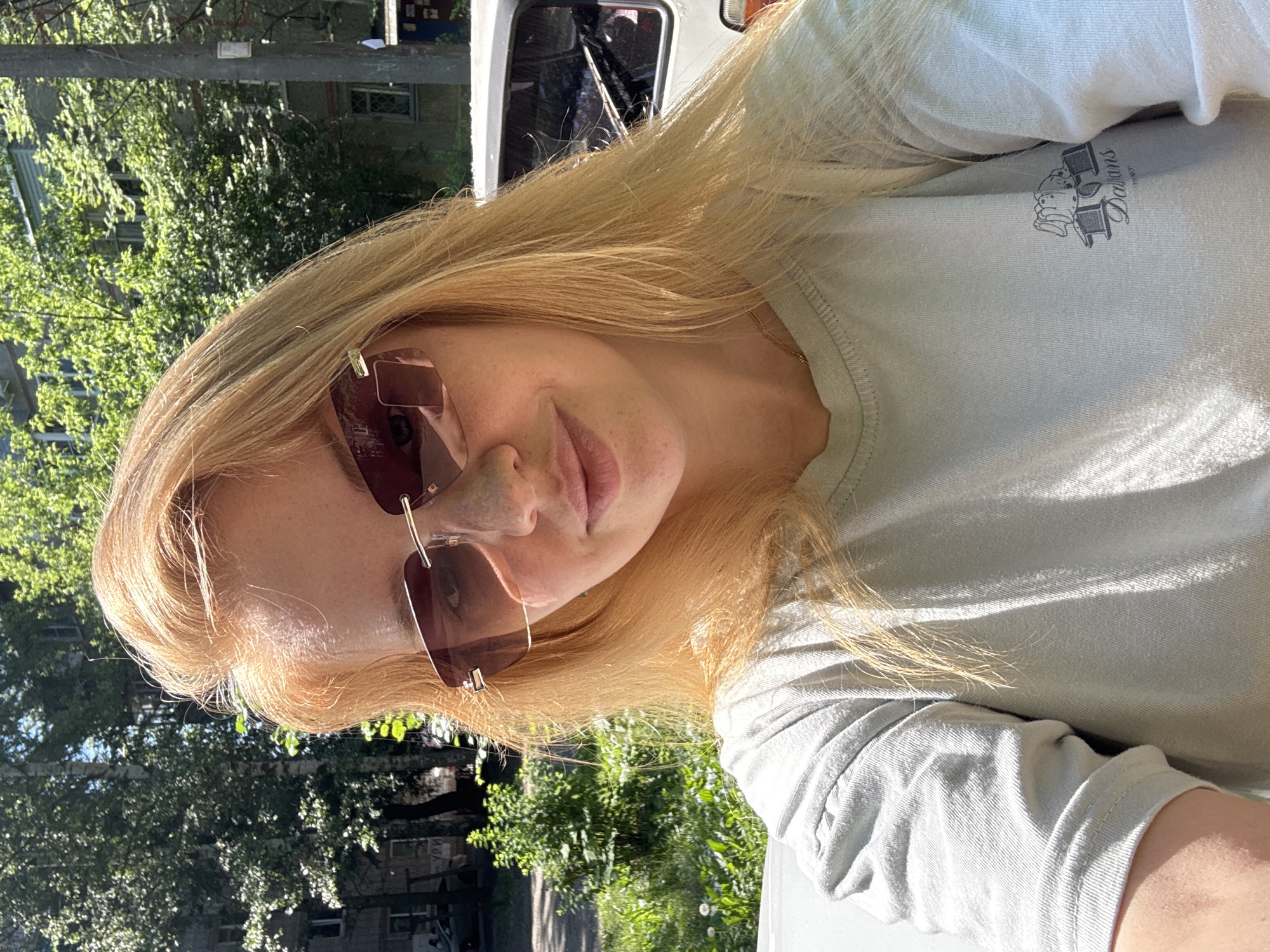 A young woman with blonde hair and sunglasses poses for a selfie outdoors on a sunny day. She is wearing a light-colored t-shirt with a logo on the right side. Her hair is parted to the side and falls loosely around her shoulders. The background shows trees, a building, and a parked car, suggesting an urban or suburban environment. The scene appears to be in the city of Yaroslavl, Russia, possibly on a street or in a park.