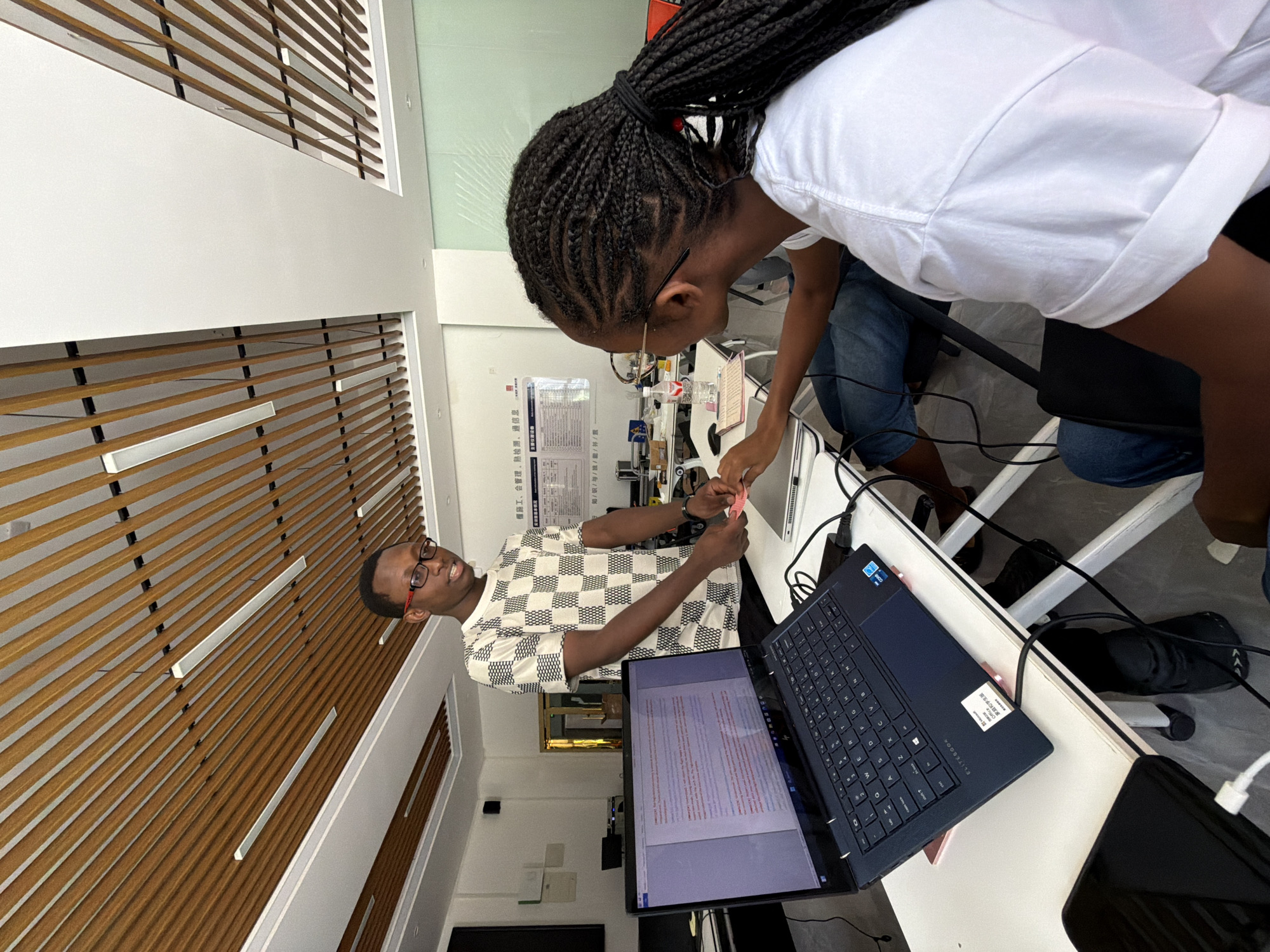 In an office setting in the city of Jiangjin, China, two individuals are engaged in collaborative work. A young Black man wearing glasses and a checkered shirt stands near a desk, holding a measuring tape in his right hand and interacting with a laptop. Facing away from the camera, a young Black woman with braided hair, wearing glasses and a white t-shirt, sits at the desk. The laptop's screen displays text, suggesting they are reviewing or working on a document. A second laptop is also visible on the desk, along with a water bottle and other office supplies. The room features a modern ceiling with wooden slats and integrated lighting, and a whiteboard with Chinese text is mounted on the wall in the background. The overall scene depicts a focused and productive work environment.