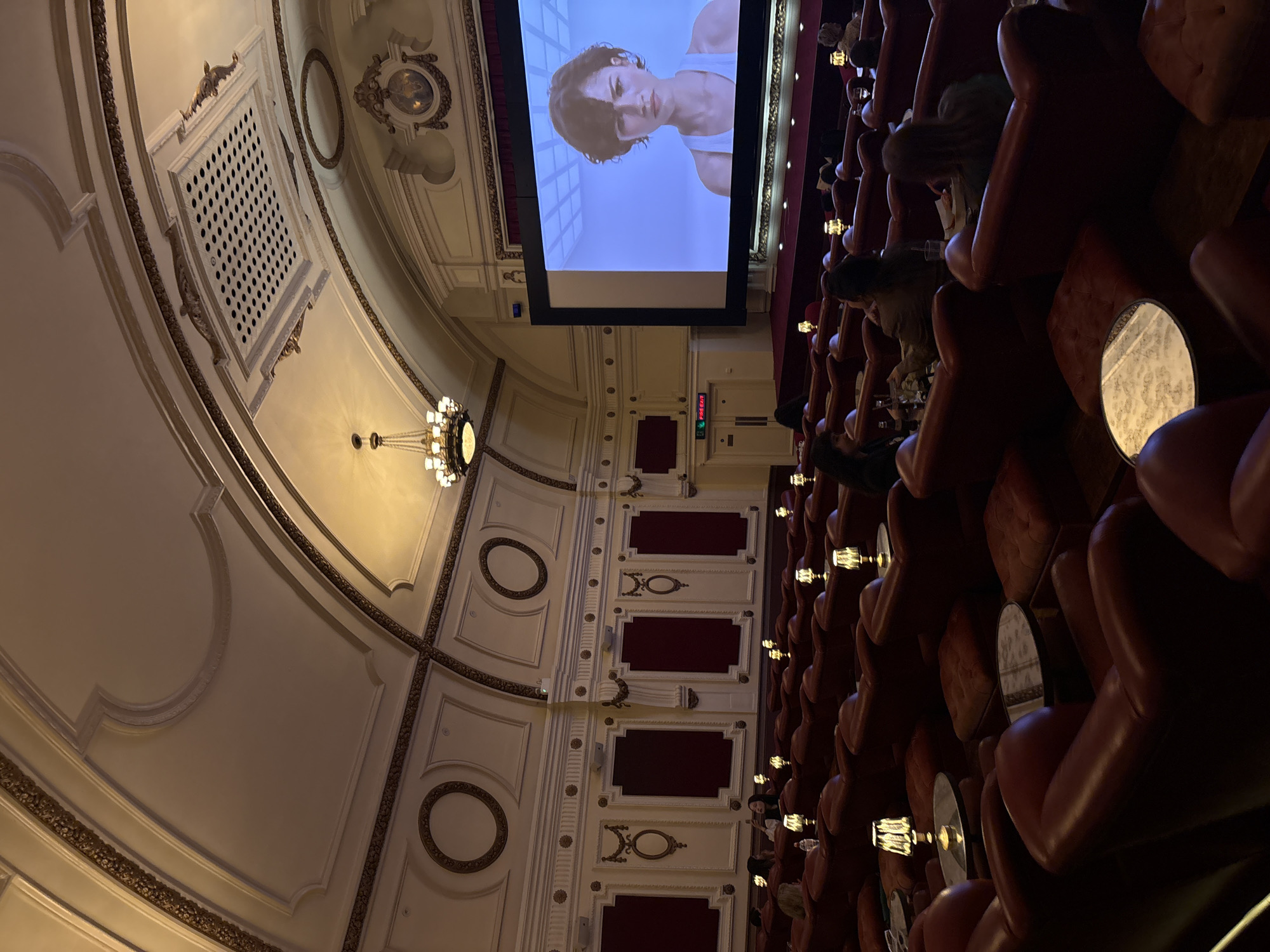 The image shows the interior of a theater, possibly located in an unknown city. The theater features a large screen displaying a video of a person, rows of plush red seats with small tables, and ornate architectural details including a decorative ceiling, ventilation grates, and classical-style embellishments. The theater is occupied by people watching the screen.