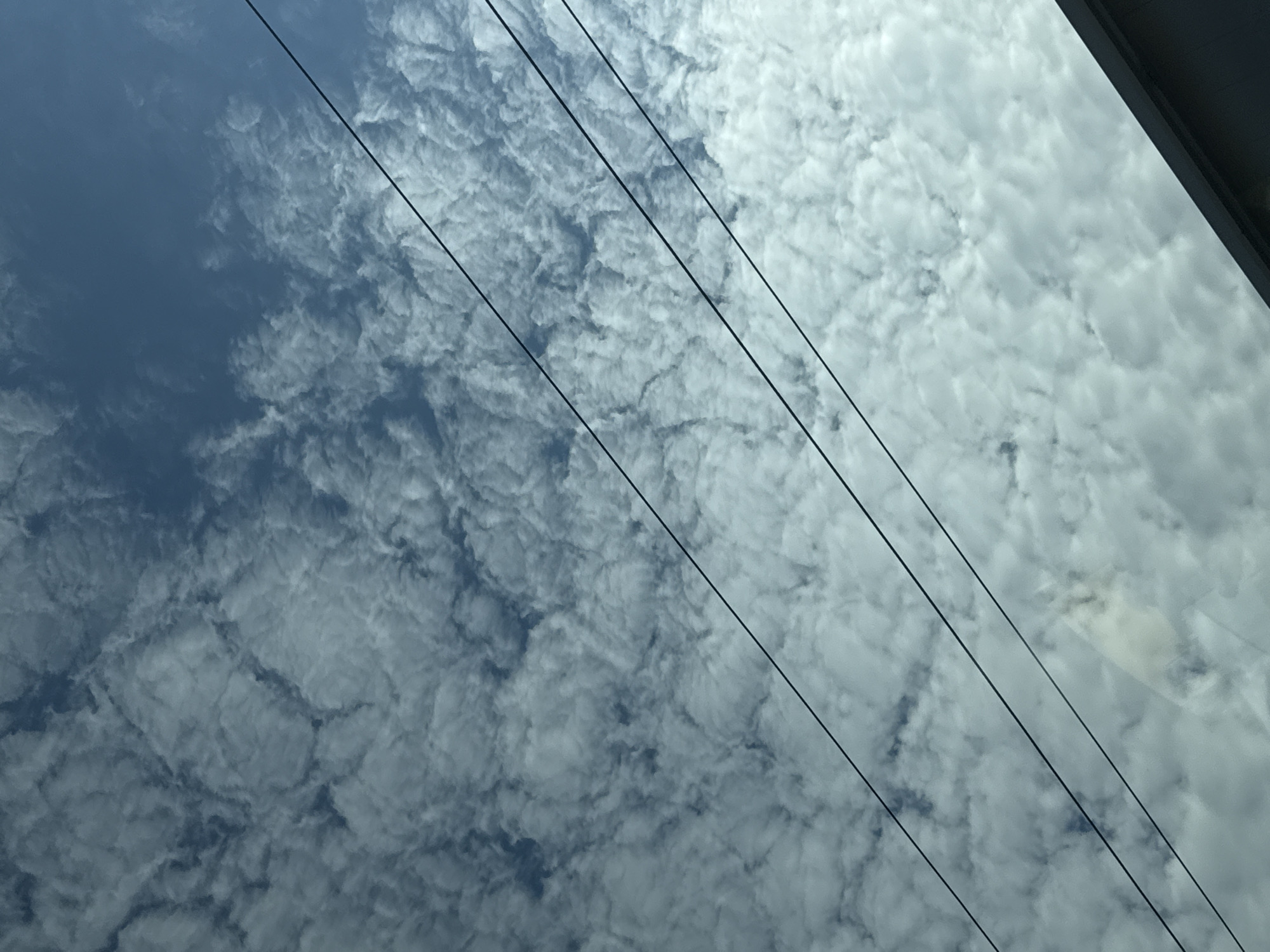 The image captures a close-up view of a cloudy sky with several parallel power lines running diagonally across the frame. The clouds are light and textured, with patches of darker blue sky visible through them. The bottom right corner of the image shows a dark, angled structure, possibly a rooftop or overhang, suggesting the photo was taken from a low vantage point, looking upwards. The scene appears to be an outdoor environment within the city of Varna, Bulgaria, characterized by the urban infrastructure of power lines set against a natural sky. There are no people or animals visible in the frame.