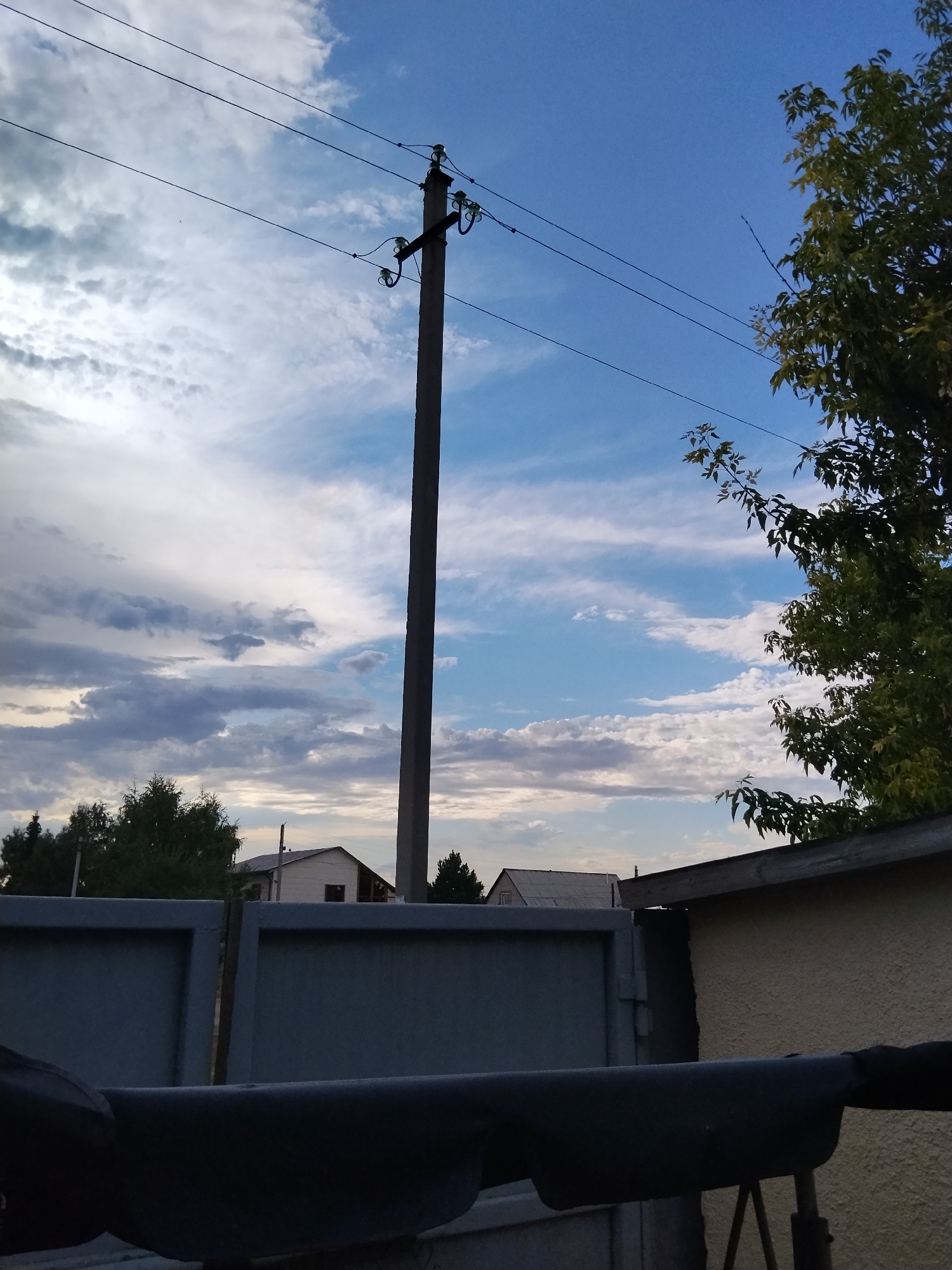 The image depicts an outdoor scene with a concrete utility pole as the central element, supporting several wires that stretch across the frame. The pole is positioned in front of a sky filled with a mix of blue and scattered clouds. To the right, a portion of a tree with green leaves is visible, partially obscuring the sky. In the background, several houses with tiled roofs and trees are visible, suggesting a residential area. A gray metal gate occupies the foreground, and a darker structure, possibly part of a fence or bench, is in the immediate foreground. The overall impression is that of a suburban or rural setting under a cloudy sky. Given the prompt's request for a specific location in Kokshetau, Kazakhstan, this image shows elements consistent with such a location, including utility infrastructure, residential buildings, and vegetation, typical of a city's outskirts.