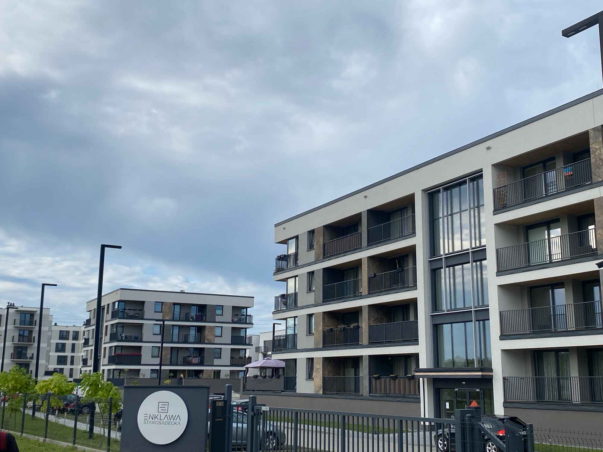 The image captures a modern apartment complex in the city of Stary Sącz, Poland. The architecture features multiple stories with balconies and large windows. The buildings are light-colored with dark accents, and the sky above is overcast with gray clouds. In the foreground, a black metal fence encloses the property, and a sign with a logo and text "ENKLAWA STAROSĄDECKA" is visible. Several black lampposts are positioned throughout the scene. There are no visible people in the image. The environment appears to be residential, with a hint of greenery from trees along the fence line.
