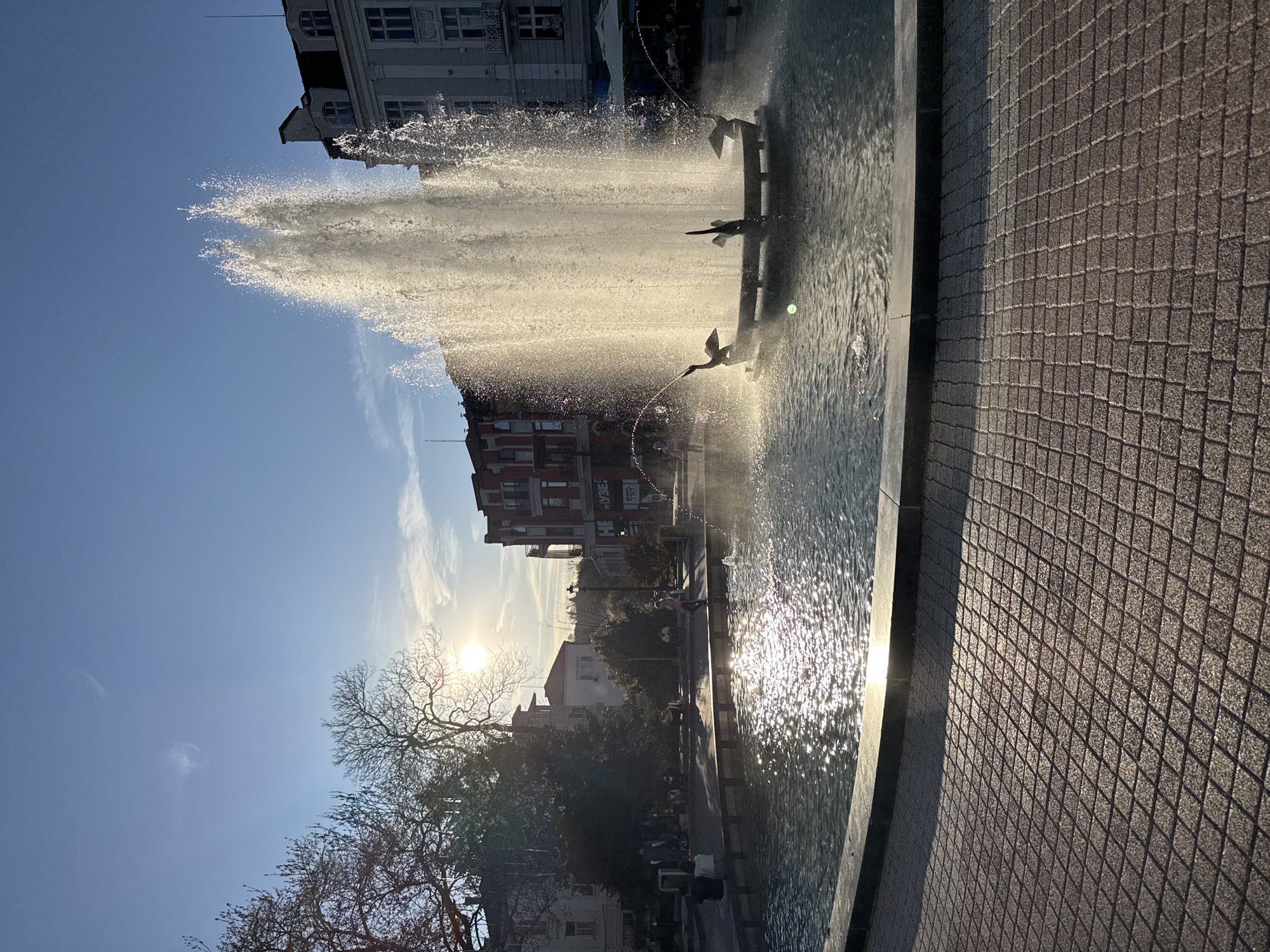 The image captures the Singing Fountains in Plovdiv, Bulgaria. A large water fountain dominates the scene, with water jets reaching high into the air. The jets reflect the sunlight, creating a sparkling effect. Sculptural elements, resembling birds, are positioned within the fountain's structure. Several buildings surround the fountain and are visible in the background. Trees are visible on the lower-left, partially obscuring some buildings. The paved area next to the fountain features a textured, geometric pattern of a repeating square. Some people are vaguely visible in the background, suggesting activity around the fountain. The sky is blue with some scattered clouds. The low angle of the sun indicates either morning or late afternoon.