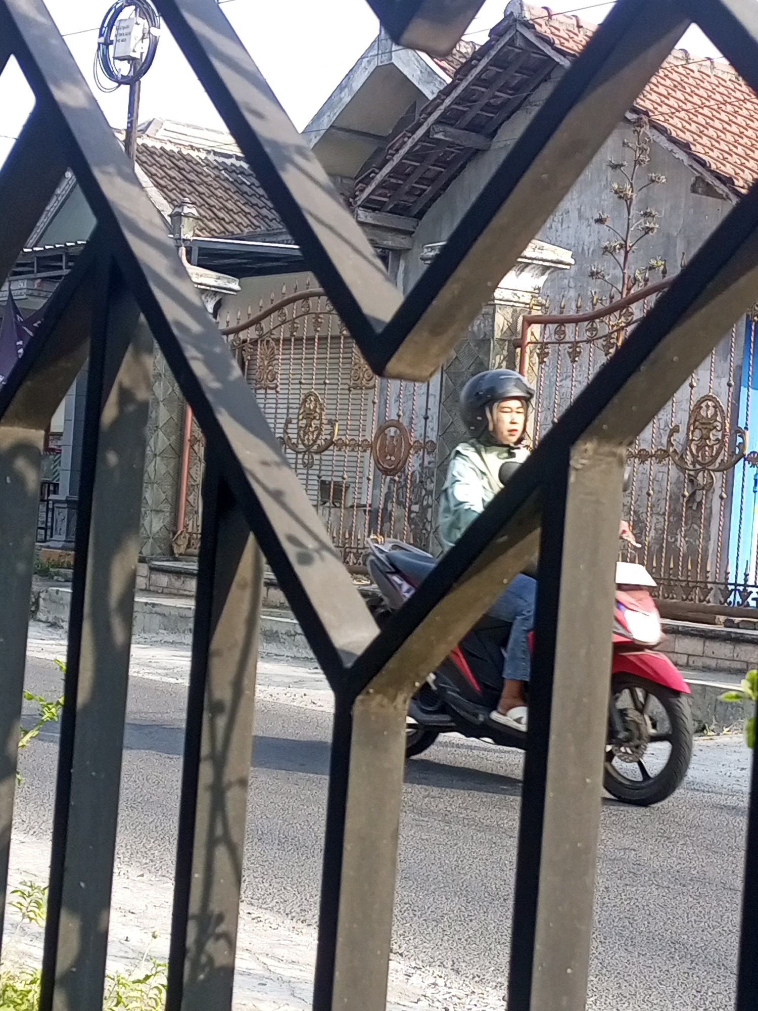 A person wearing a black helmet and a light green jacket is riding a red and black motorcycle on a paved road. The person has their eyes closed. The motorcycle has a silver front fender and chrome accents. The background consists of a house with a tiled roof and a decorative metal fence. A utility pole with wires is visible to the left. The scene is illuminated by sunlight, casting shadows on the road. This image was captured in Ngasem, Indonesia.