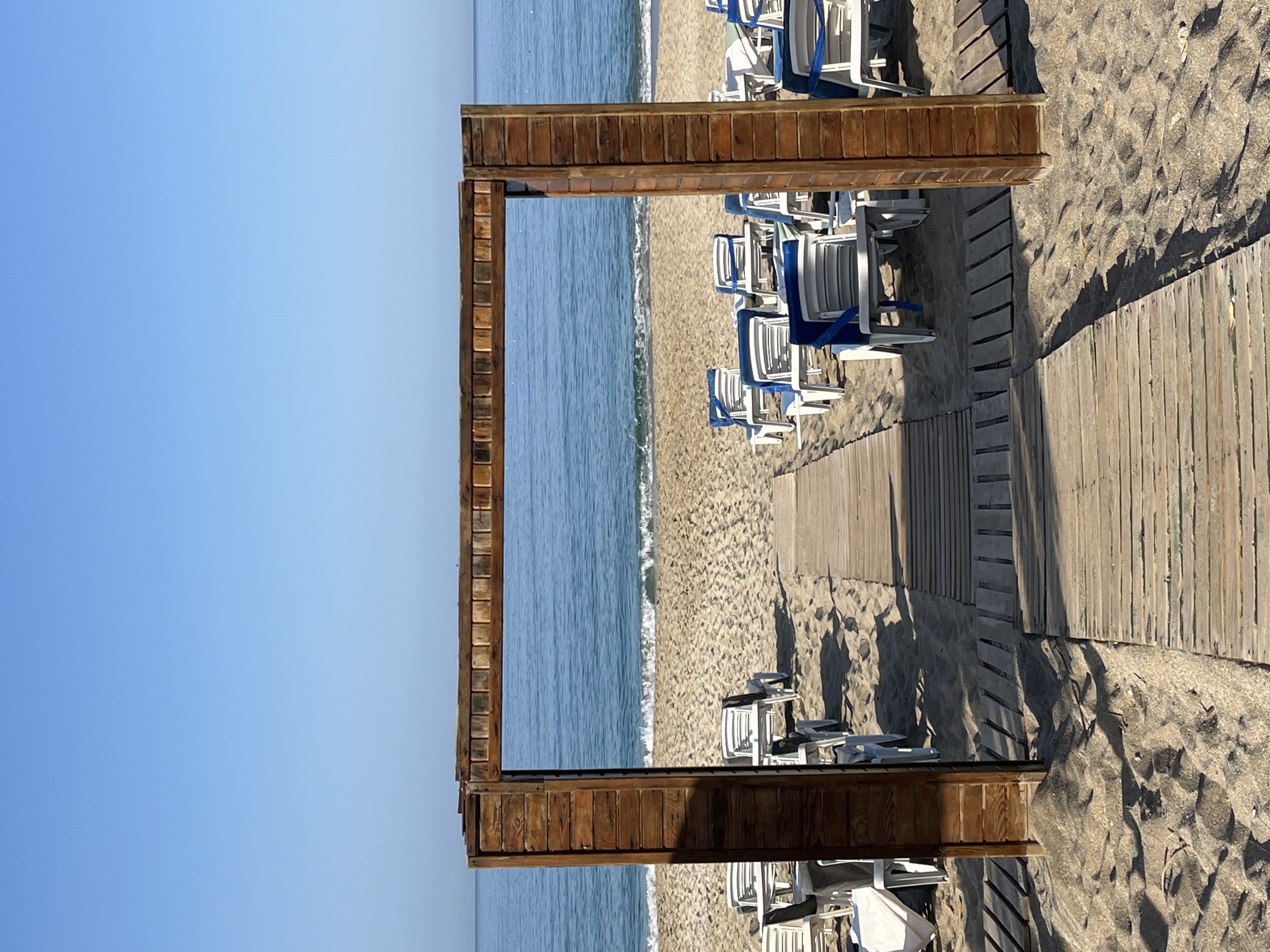 The image depicts a beach scene, likely in Alanya, Turkey, judging by the beach chairs and wooden structure. A wooden frame stands perpendicular to the shoreline. The sea is visible through the frame, transitioning into the clear blue sky. To the right of the frame is a beach with white and blue beach chairs. A wooden walkway stretches across the sand. The sand shows patterns of footprints and shadows.