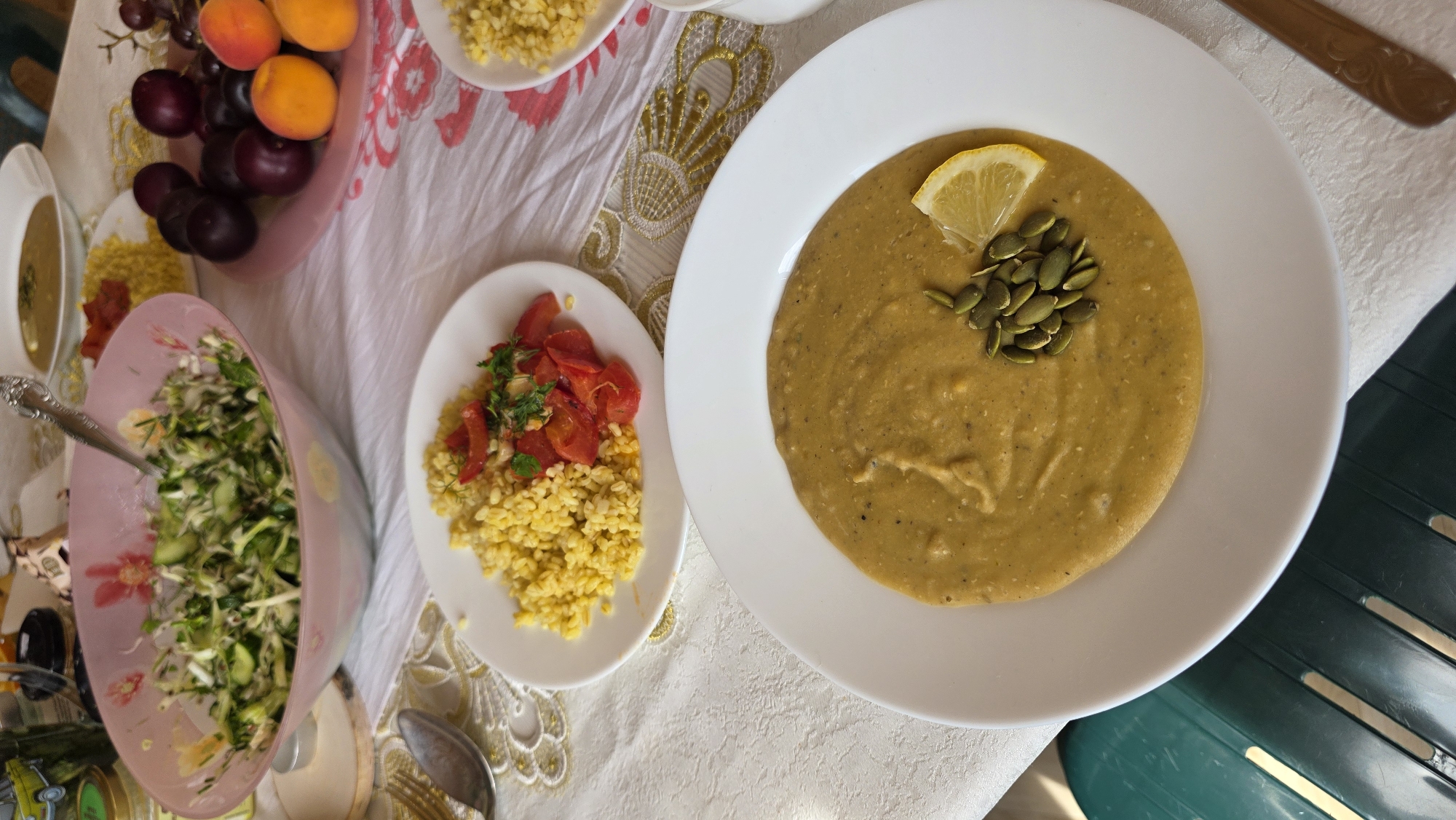 A table is set with a meal, featuring several dishes. In the foreground, a white plate holds a bowl of golden-hued soup, garnished with pumpkin seeds and a lemon wedge. To the left of the soup, another white plate is visible with a portion of yellow grains topped with chopped red tomatoes and herbs. Behind these plates, a large pink bowl contains a salad composed of shredded green vegetables and herbs, with a spoon inside. Further back, a pinkish plate displays a selection of dark plums, apricots, and grapes. A white bowl of soup is also visible in the background, and a jar with a yellow lid is present on the left side of the table. The table is covered with a white tablecloth that has a decorative pattern. The scene appears to be indoors, as suggested by the lighting and the presence of a tablecloth, and is likely set in the city of Kupoyarovo, Russia, based on the provided context.