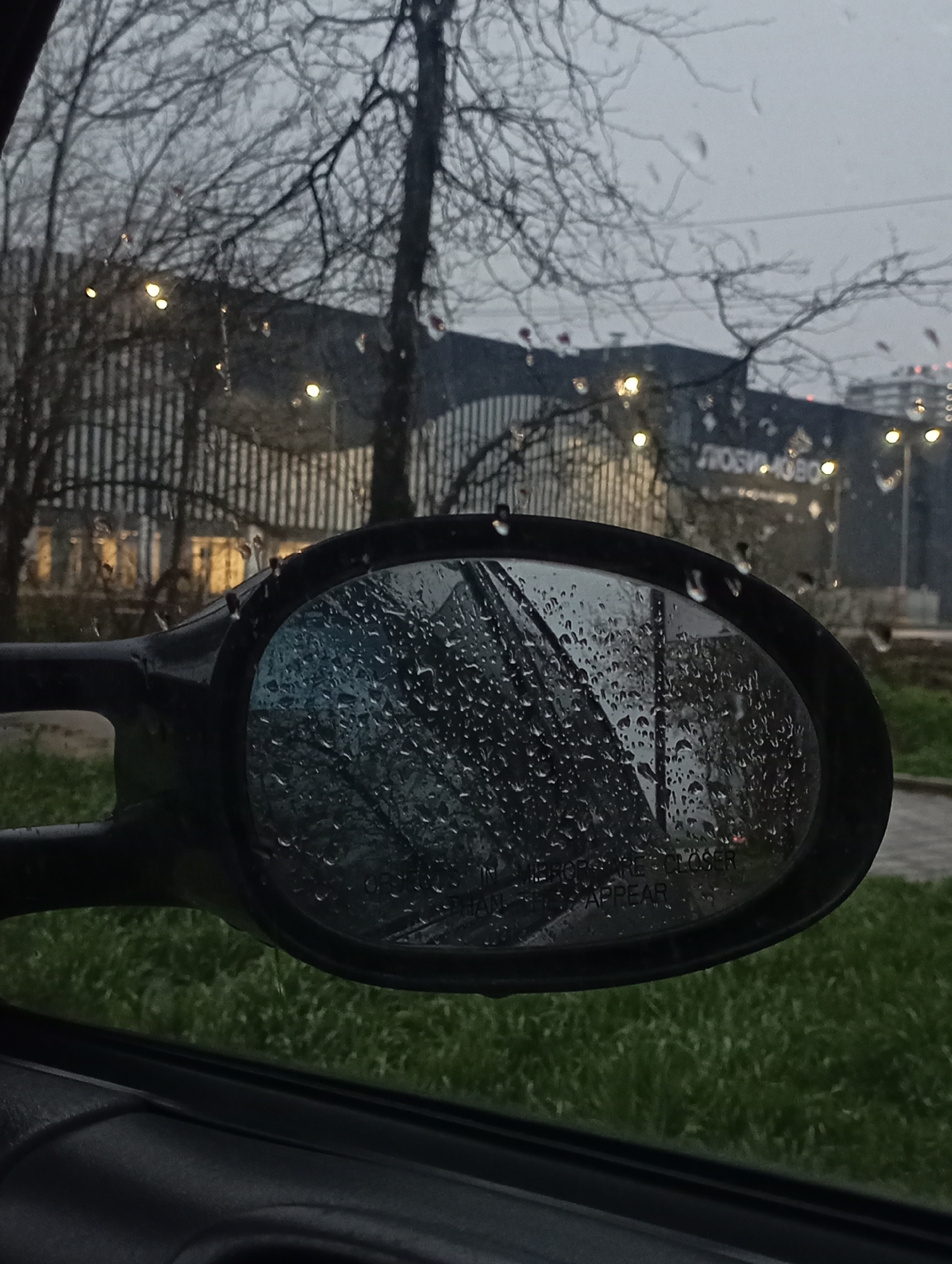 The image shows a car's side mirror covered in raindrops, reflecting a blurred view of the outside. In the background, a building with vertical lines and the words "Любимово" (Lyubimovo) are visible. Bare trees frame the scene. The environment suggests a rainy day in an urban setting. Based on the presence of the "Любимово" sign, the location is likely in Krasnodar, Russia. Green grass is visible below the side mirror, further outside the car. The interior of the car is visible at the bottom of the frame.