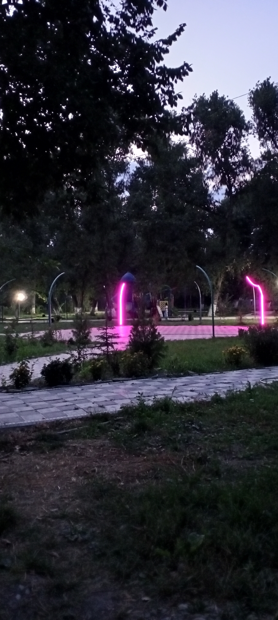 The image depicts a park area at dusk in Sokuluk, Kyrgyzstan. A paved pathway with a checkered pattern runs through the foreground, bordered by grass and some small bushes. In the mid-ground, a playground structure is visible, with two individuals standing near it. The park is illuminated by ambient lighting, including a street lamp on the left and neon pink archways that add a vibrant, artificial glow to the scene. Tall trees with dark foliage dominate the background, silhouetted against the pale, twilight sky. The overall atmosphere is serene, transitioning from day to night.
