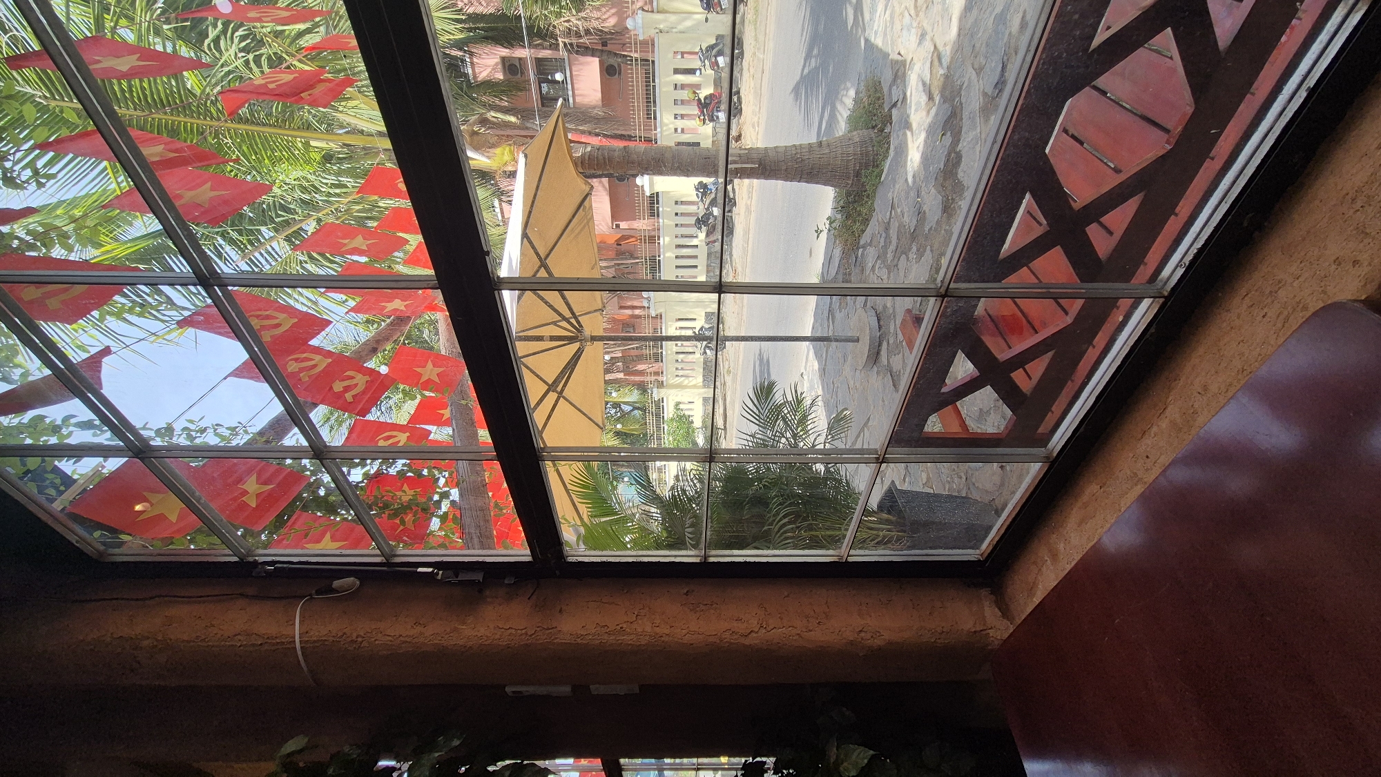 The image shows a view from inside a building looking out through a large, multi-paned window onto a street scene in Đà Nẵng, Vietnam. Red flags with yellow stars, characteristic of Vietnam, are strung across the top portion of the window. Outside, a paved walkway leads to a street. Palm trees are visible, with one mature palm tree to the right of the frame. An open, yellow umbrella stands on the walkway. Buildings with a light pink facade and white railings line the street. Several motorcycles are parked along the roadside. Inside the building, a portion of a dark reddish-brown table is in the foreground, and part of a dark brown textured wall is on the left. A potted plant with palm-like fronds is visible through the window.