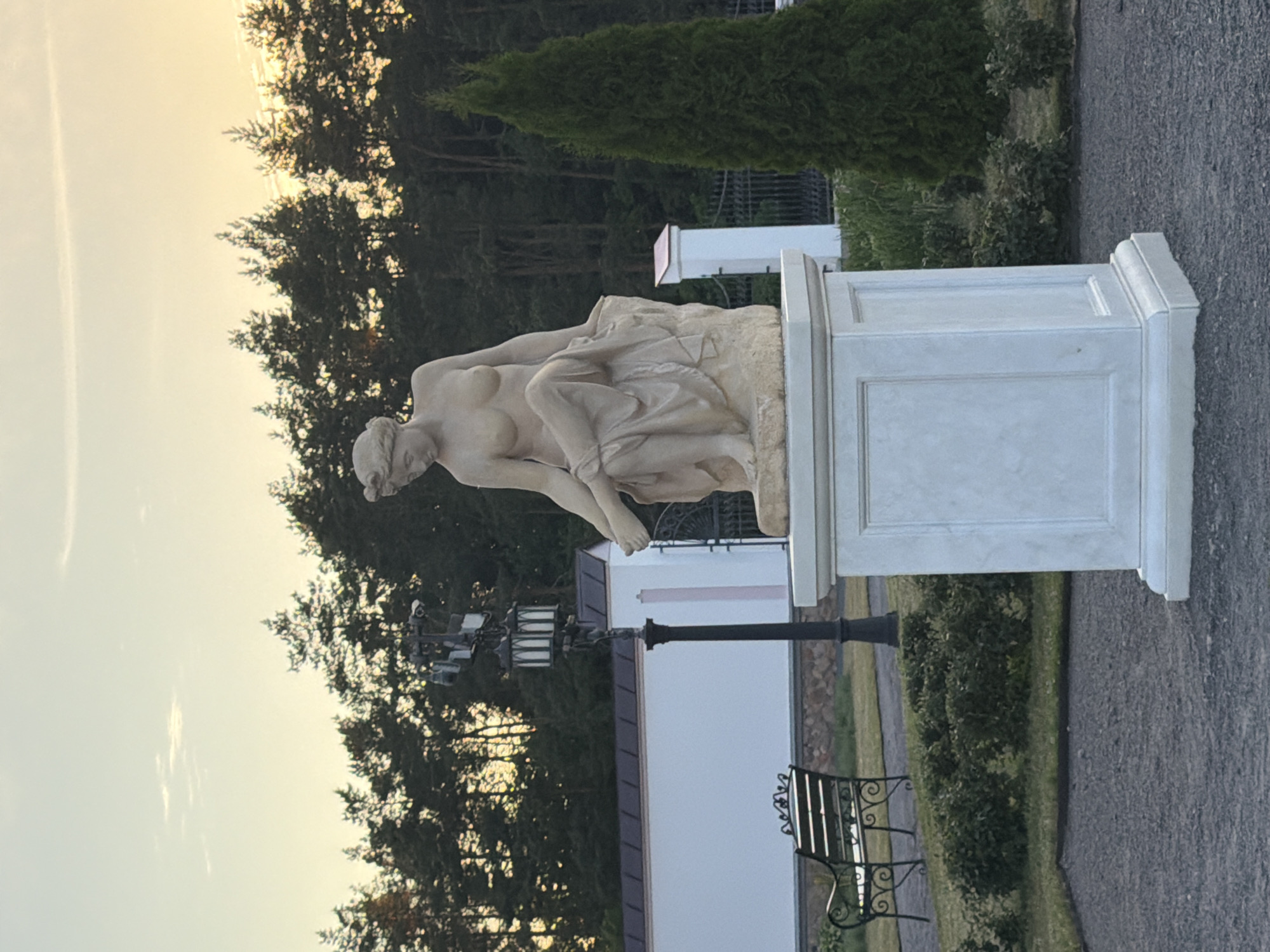 This media file shows a stone statue of a nude female figure reclining on a rock. The statue is placed on a white pedestal in what appears to be a park or garden setting. The background features lush green trees, some of which are tall evergreens, and a clear sky with a soft glow, suggesting it might be dawn or dusk. There is a white building with a dark-pitched roof visible behind the statue. A black lamppost stands in front of the building, and a wrought-iron bench is also present. The overall scene evokes a sense of tranquility and historical or artistic appreciation. The lighting suggests a calm, possibly serene atmosphere. The location is not specified.