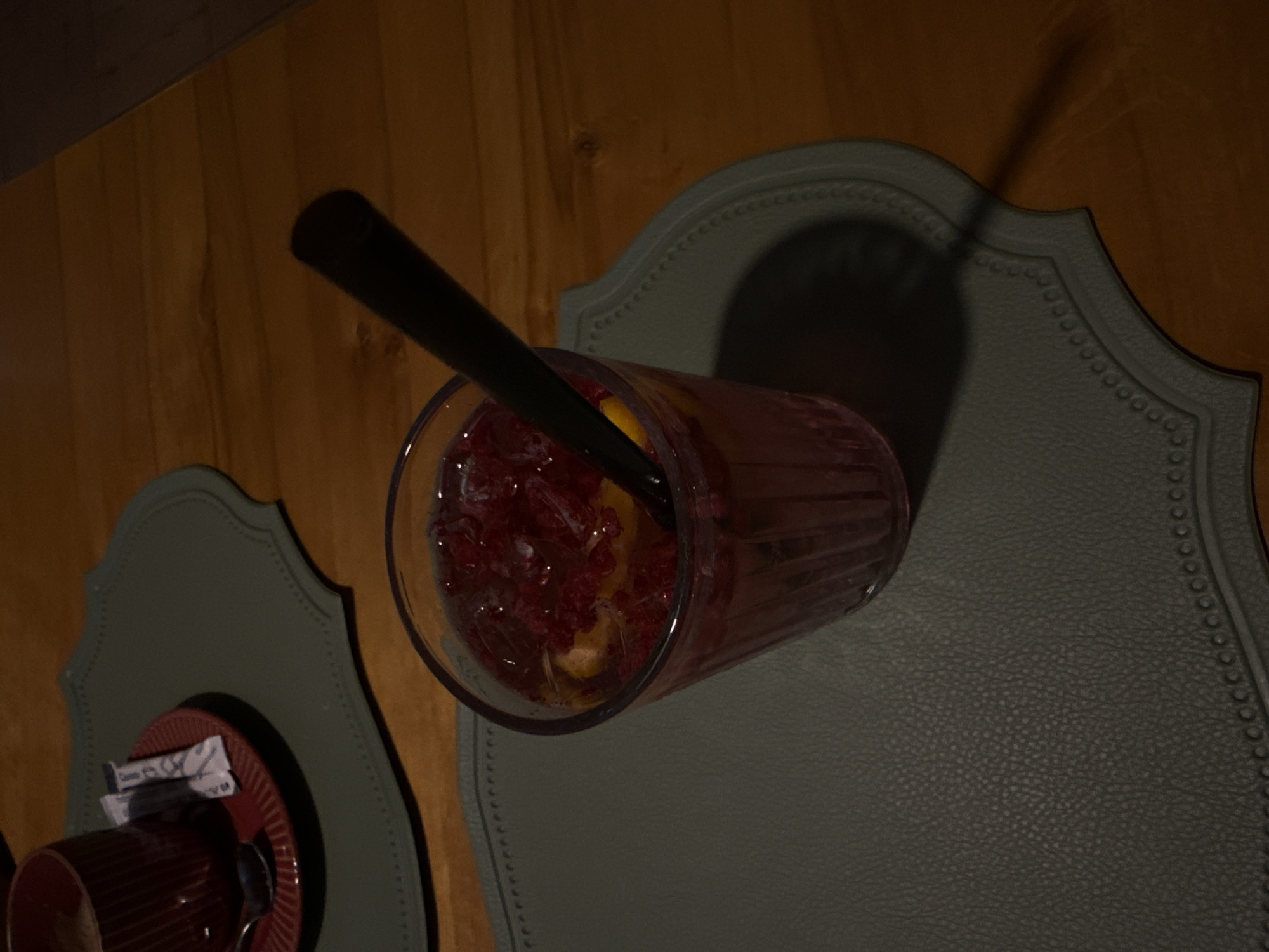 A high-angle, dimly lit shot features a glass of red liquid with raspberries and ice, a black straw, and a red cup with a spoon on a saucer with two sugar packets. The glass and cup are placed on grey placemats on a wooden table. The setting appears to be a cafe in Abinsk, Russia.