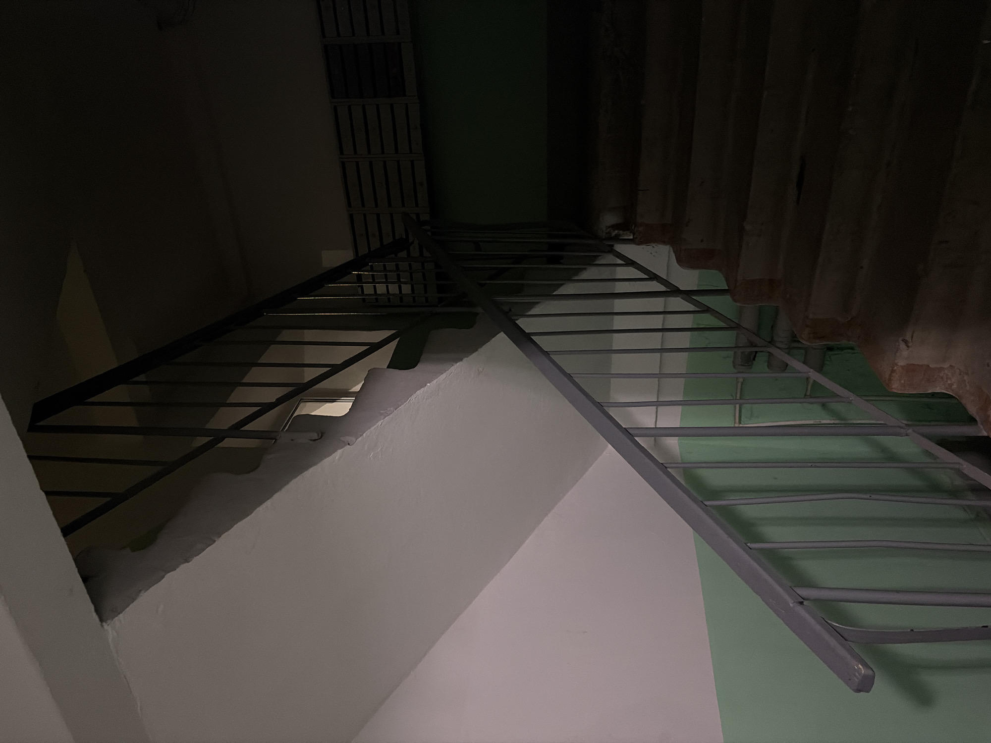 A low-light, diagonal view captures an interior staircase within what appears to be a functional or industrial section of a building in Perm, Russia. The scene is dominated by two sets of unadorned, dark gray metal stairs and railings. One flight ascends diagonally from the lower left, while another, more horizontal, is visible in the upper right, suggesting a landing or continuation of the ascent.
The walls are painted in a stark combination of muted light gray and a pale, dull green. In the upper left, a dark, vertical structure composed of slats or grilles is visible, potentially a ventilation shaft or pipe enclosure. The upper right corner reveals a dark, corrugated ceiling or wall surface, adding to the utilitarian aesthetic.
The lighting is notably dim, casting deep shadows across the structures and contributing to a somber or stark atmosphere. No individuals are present, and no activity is occurring. The lack of discernible natural light indicates either a windowless space or nighttime conditions. The practical design of the stairs and the muted color scheme evoke a sense of an older, functional building. No text is visible in the image.