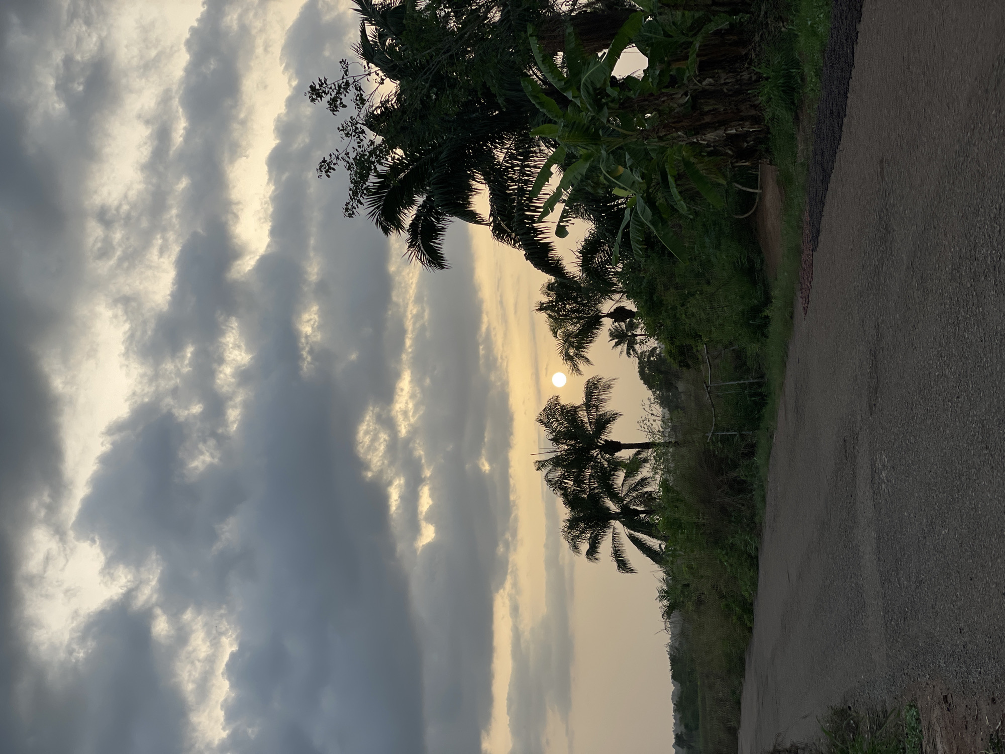 The image depicts a road in Agba Umana, Nigeria, at dusk or dawn. The sky is filled with dark, textured clouds, and the sun is visible, partially obscured by the clouds. Silhouetted palm trees and other vegetation line the side of the road. The road itself is paved and appears to be slightly damp. There are no visible people or other subjects other than the natural elements.