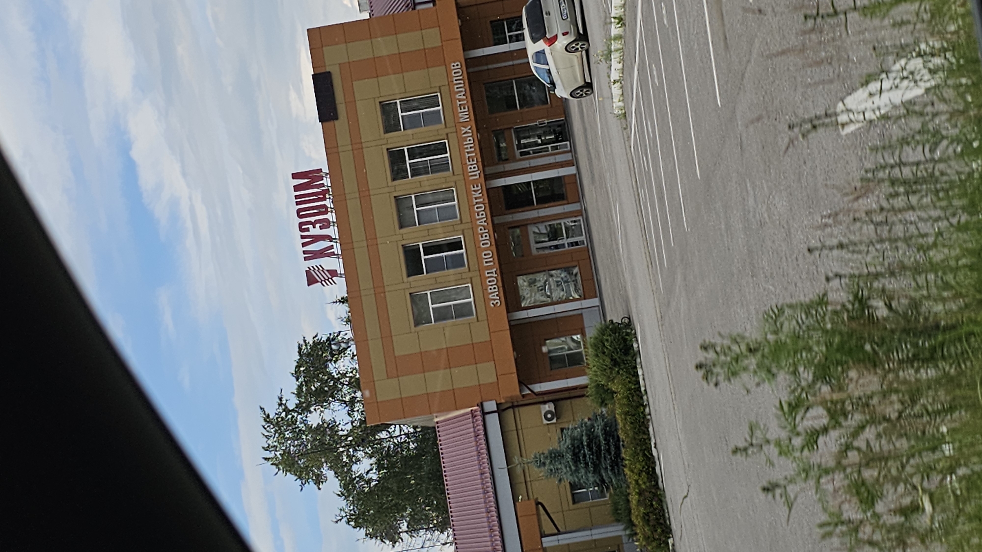 The image shows the facade of a building with a sign that reads "КУЗОЦМ" in red letters. Below this sign, another sign states "ЗАВОД ПО ОБРАБОТКЕ ЦВЕТНЫХ МЕТАЛЛОВ" in white letters. The building has an orange and brown patterned exterior and several windows. To the left of the main building, there is a smaller yellow structure with a maroon roof. In front of the building, there is a parking lot with white lines marking parking spaces. A white car is parked in the lot on the right side of the image. There are green trees and bushes in front of the buildings and tall grass in the foreground. The sky is blue with white clouds. The overall scene appears to be an industrial or commercial property. This is likely the "KUZOCM" plant, which is located in Kamensk-Uralsky, Russia, and is known for processing non-ferrous metals.
