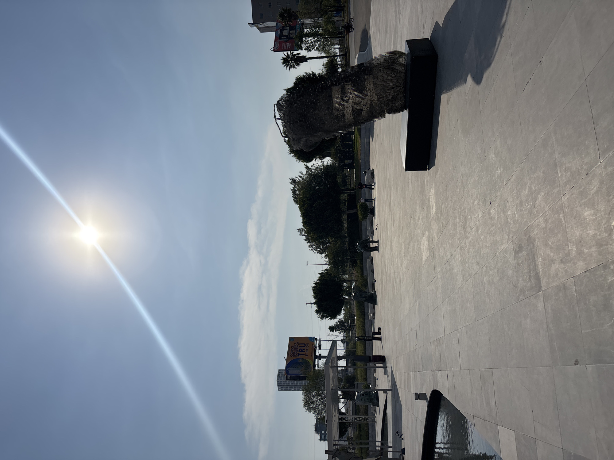 The image depicts a sunlit outdoor scene in San Bernardino Tlaxcalancingo, Mexico. A large, abstract wire sculpture of a human head is prominently featured. The sculpture is placed on a dark-colored rectangular base. The setting appears to be a public space or park, evidenced by the paved ground, trees, and other sculptures visible in the background. There are some people visible in the distance. A large billboard advertisement is visible.