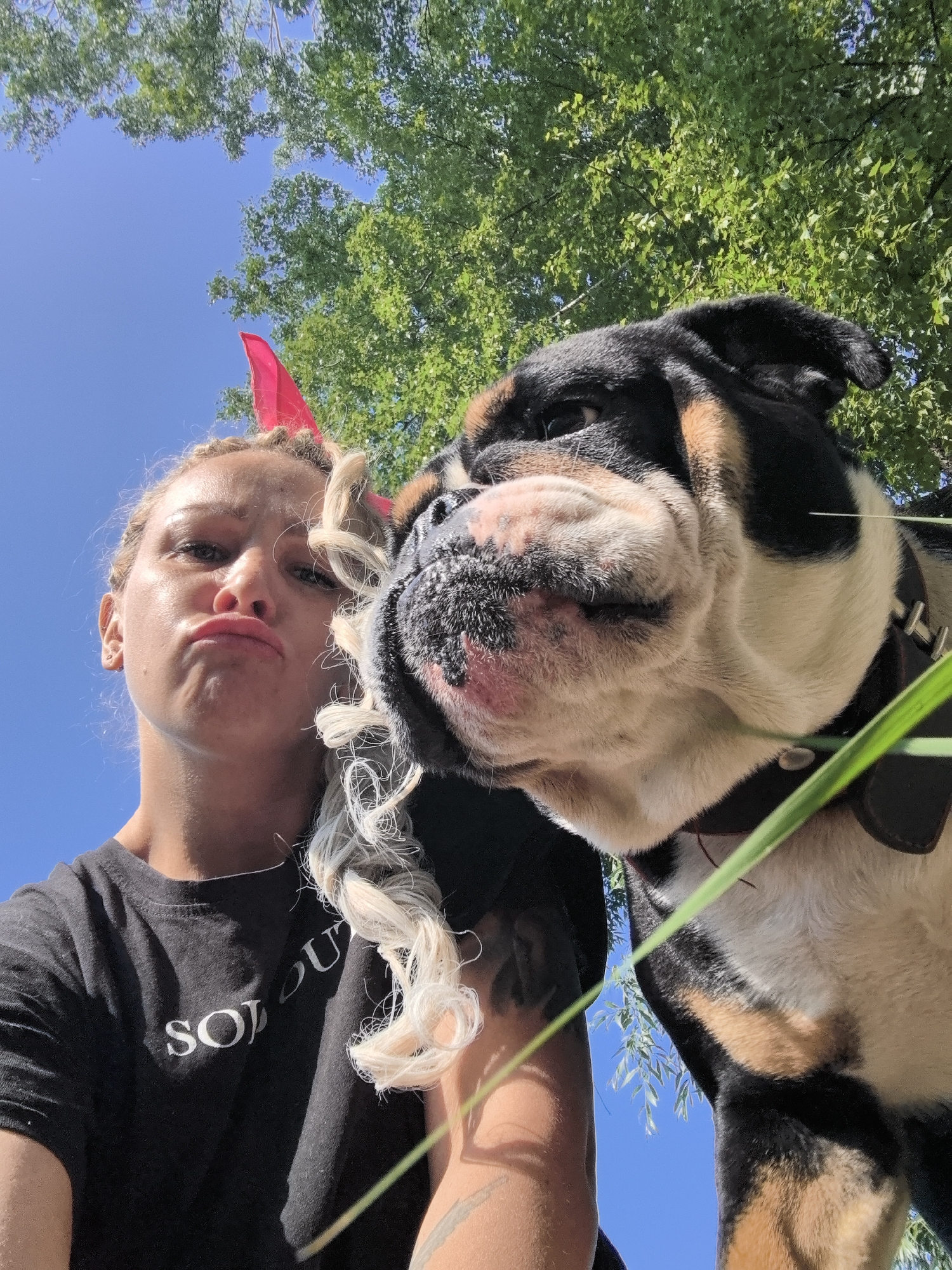 An eye-level, low-angle, outdoor, full-body, selfie-style shot features a person and a dog with a clear blue sky and green trees in the background. The person, positioned on the left, has blonde hair styled in braids with curly strands framing their face, and appears to be wearing red devil horns. They are looking directly at the camera with a pouty expression, and are wearing a black t-shirt with white lettering that reads "SOLO U." The dog, an English bulldog, is on the right, with its face close to the person's face. The dog has black, tan, and white markings, a dark collar, and is looking slightly to its left. The overall scene suggests a playful moment captured outdoors, possibly in a park or backyard, given the natural environment. The image does not provide any specific information about the location within the city of Noviy, Russia.