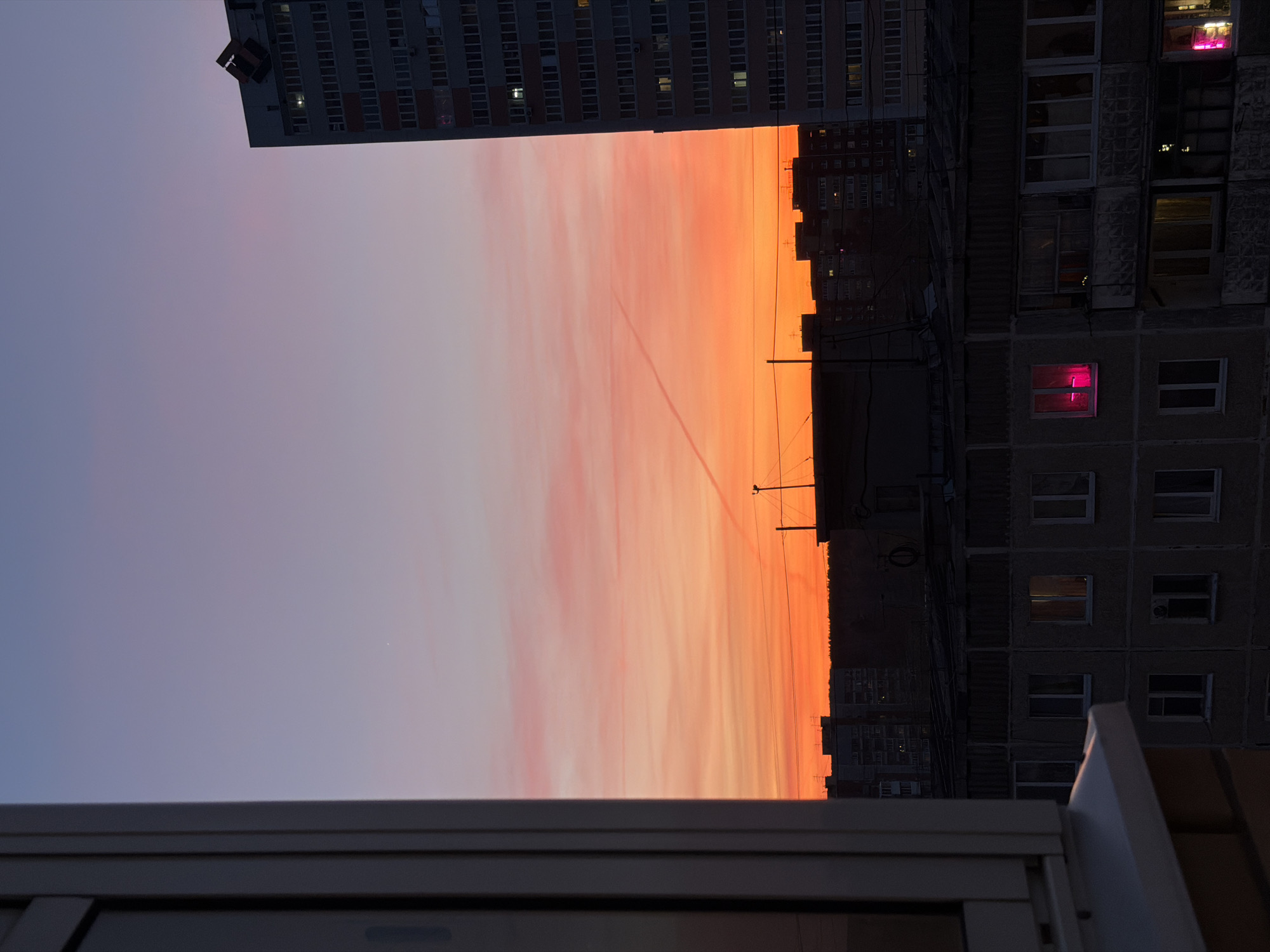This media file captures an urban twilight scene, likely a sunset, viewed from a window or balcony. The image is composed of three main horizontal bands: a silhouetted tall building at the top, a vibrant sky in the middle, and a window frame in the foreground at the bottom.
In the top section, a modern, multi-story residential building extends across the frame, its many rectangular windows showing faint internal lights. To the right, an older-style, blocky residential building with a textured facade rises vertically, its windows darker save for one striking exception: a window emitting a bright, intense magenta or pink glow.
The central and most prominent feature is the sky, which displays a dramatic color gradient. It transitions from a soft blue-purple at the top to fiery oranges and pinks towards the bottom, indicative of dusk or dawn. Long, streaky clouds are visible within the warmer hues, and a thin, straight contrail or vapor trail cuts diagonally across this colorful expanse. Faint power lines are also visible against the brighter part of the sky.
The foreground shows parts of a grey and white window frame or balcony railing, framing the urban vista. No people or discernible text are present. The scene evokes a tranquil yet striking moment, contrasting the natural beauty of the sky with the starkness of urban architecture, with the vibrant pink window adding an unexpected splash of color. The architectural style aligns with urban environments found in Perm, Russia.