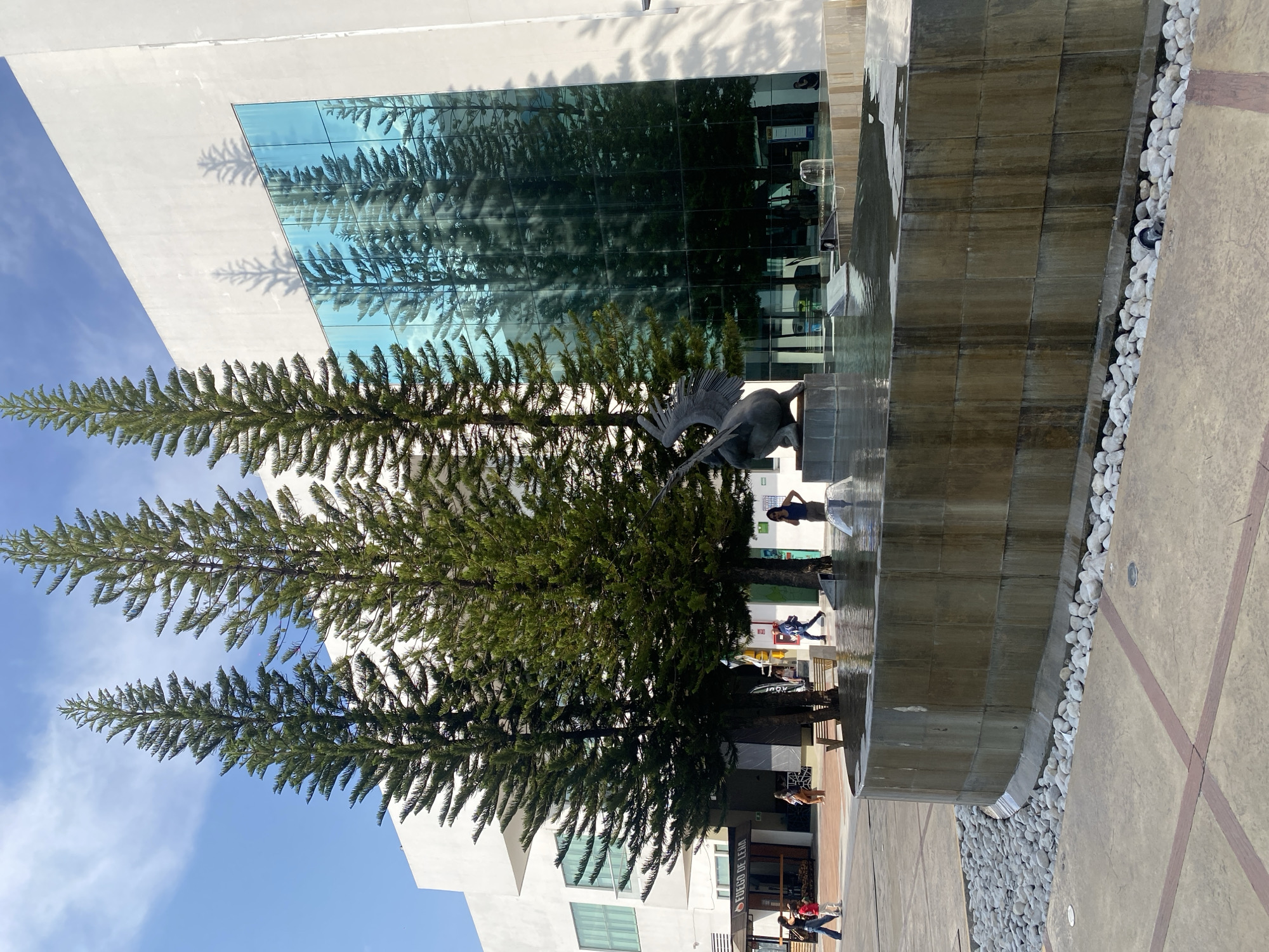 Here is a detailed description of the image:
**General Description:**
The image captures an outdoor scene with a blend of nature and architecture. The setting appears to be a modern urban space, possibly a plaza or courtyard.
**Environment and Architecture:**
* **Buildings:** A contemporary building features prominently in the background, characterized by a glass facade that reflects the surrounding trees. The building has a clean, modern design with angular edges.
* **Fountain:** A water feature, possibly a fountain or a cascade, stands near the building. The structure is built of stone or tile and is partially covered in white gravel.
* **Trees:** Several tall, slender trees with dense foliage are visible. Their positioning is such that they frame the building and fountain, adding a natural element to the urban setting.
**Objects and Subjects:**
* **Statue:** There is a statue near the fountain.
* **People:** Several people are walking in the background, suggesting that the location is accessible to the public.
**Overall Impression:**
The image conveys a sense of modern urban design integrated with natural elements. The reflection in the building’s glass facade creates a visual effect, merging the architecture with the surrounding trees.
The location is most likely the city of Unknown, Unknown.