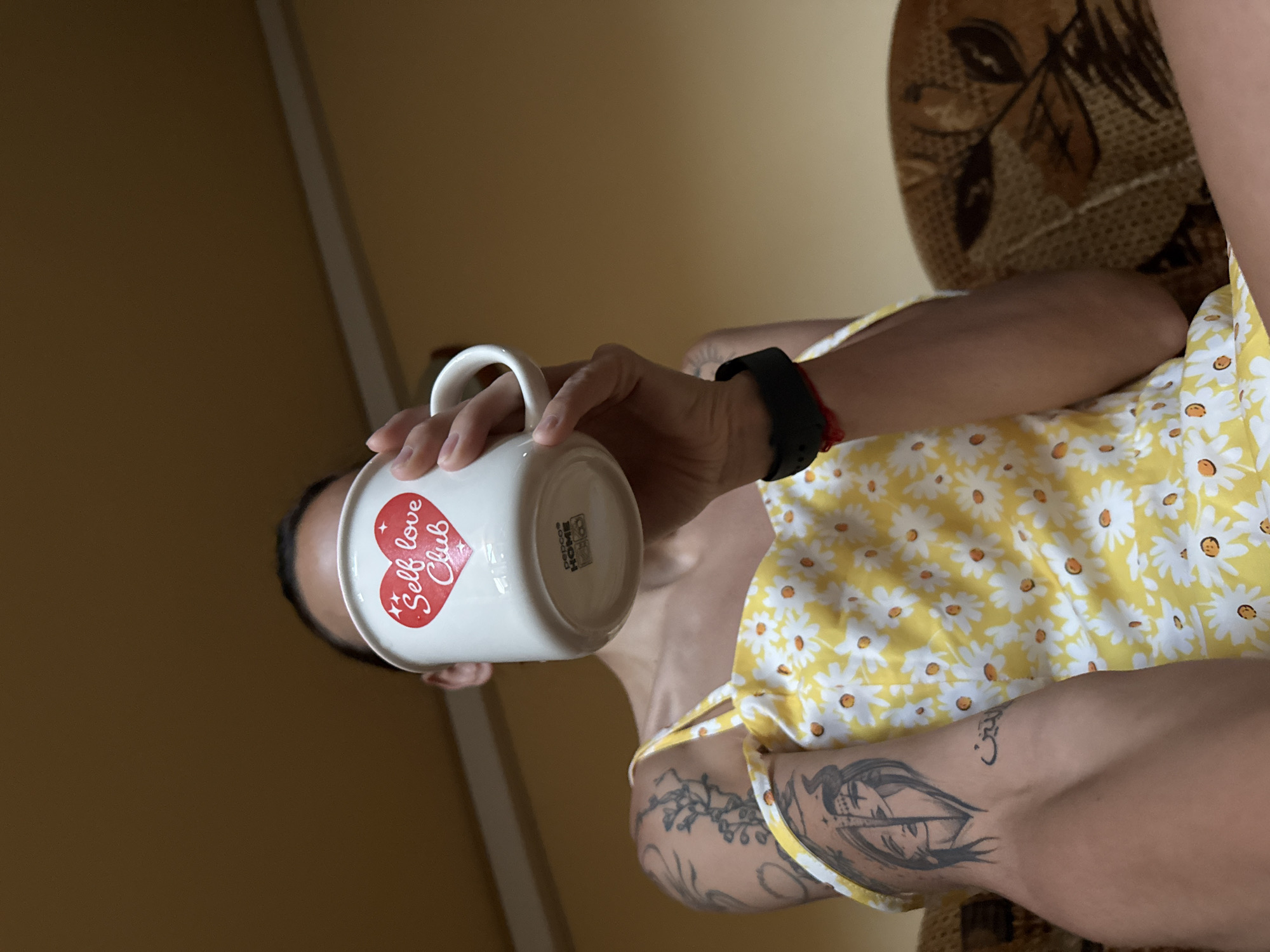The image shows a person holding a mug with the text "Self love Club" written on it, partially obscuring their face. The person is wearing a yellow dress with a daisy pattern and has tattoos on their arm. The background appears to be indoors, with a brown ceiling and a portion of a patterned sofa visible. The image does not provide any information about the location in the city of Pleven, Bulgaria.