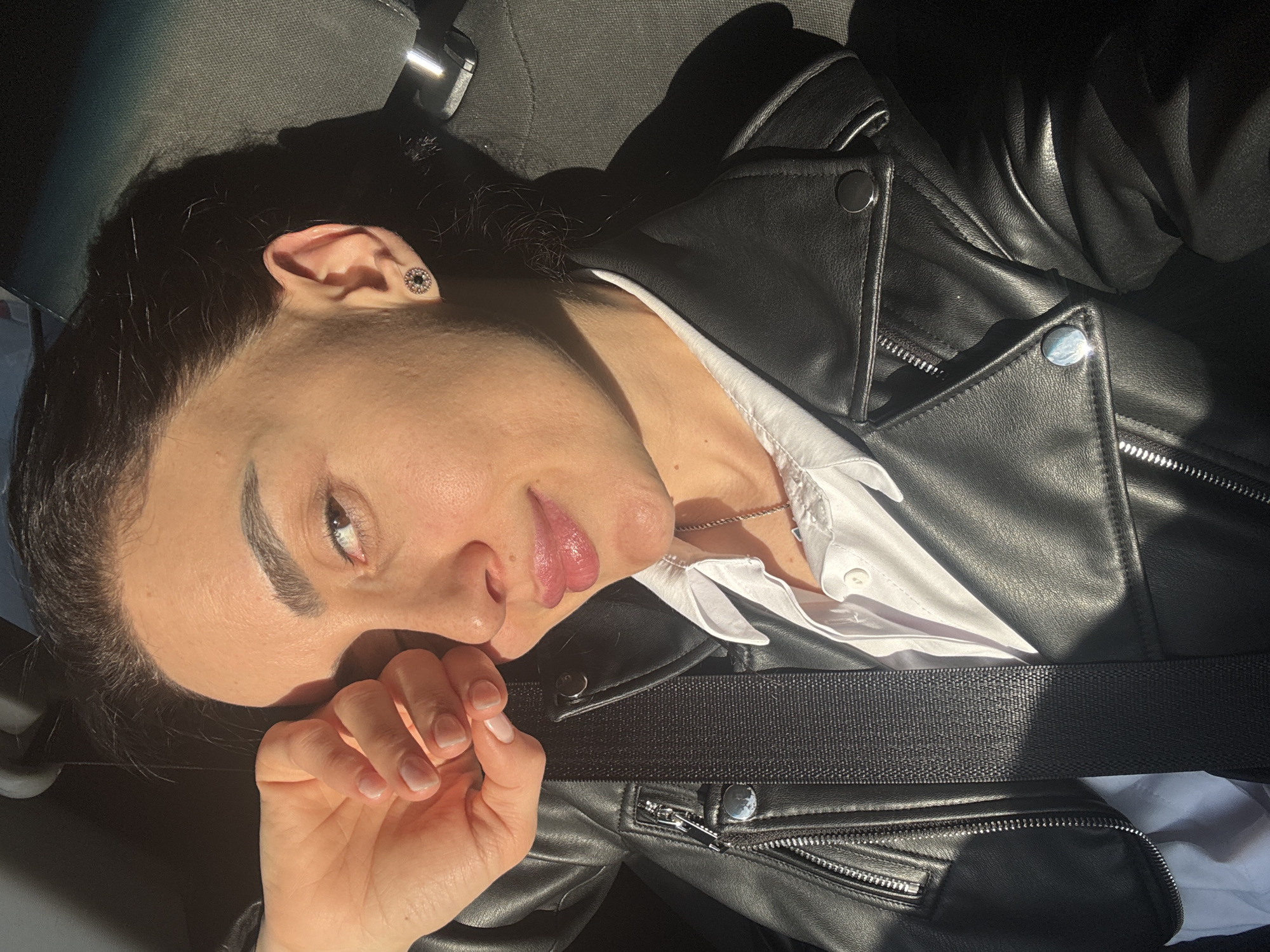A medium shot shows a light-skinned woman lying at an angle in what appears to be a car. She is wearing a black leather jacket and a white button-down shirt. A black seatbelt crosses her torso. She wears a small earring in her left ear and a delicate necklace. Her black hair is partially shadowed. Her right hand is near her face. The lighting suggests it is daytime, with sunlight entering the car. There is no indication of the location being Tbilisi, Georgia, based solely on the visual information provided.