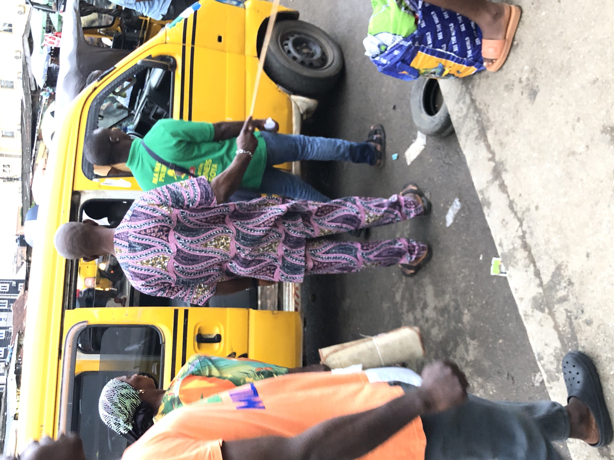 The image depicts a street scene in Lagos, Nigeria, with a bright yellow commercial bus as the central element. Several people are visible around the bus, suggesting a busy urban environment, possibly a bus stop or a market area.
In the foreground, a person wearing an orange t-shirt and grey pants is partially visible, with their left arm and hand prominent. To their right, a woman wearing a headscarf with a black and white pattern and a colorful patterned top stands near the bus door. Next to her, a man in a pink and purple patterned traditional African outfit, with a matching head covering, stands facing away from the camera. He appears to be interacting with someone to his right, possibly gesturing with his right hand.
To the right of the man in the patterned outfit, another man is seen from the back, wearing a bright green t-shirt with writing on it and blue jeans. He is standing near the driver's side of the bus. A portion of a tire and a colorful bag are on the ground in the lower right corner of the image.
The environment suggests a typical Lagos street, characterized by the presence of commercial vehicles, pedestrians, and the general hustle and bustle of city life. The buildings in the background, though blurred, hint at the dense urban architecture found in Lagos. The overall impression is one of everyday activity and movement in a Nigerian metropolis.