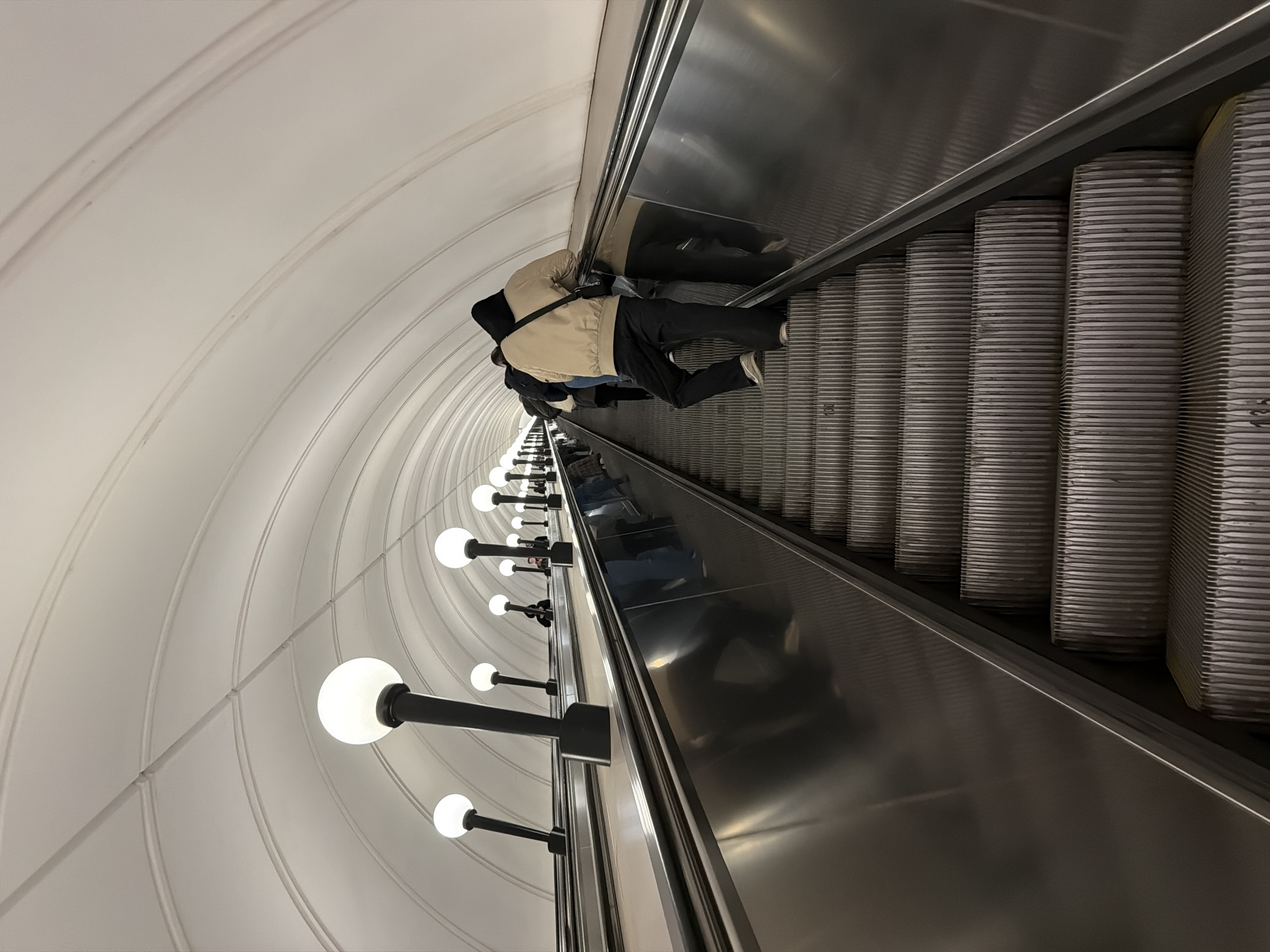 A low-angle, indoor shot shows a receding escalator in a tunnel with several people riding downward. The environment includes a rounded tunnel with white tile and rows of globular lights fixed along the left side. The escalator has metal treads and a smooth metal side. A person closest to the camera is wearing a tan jacket, dark pants, and white sneakers. The people are going down the escalator. The location is an underground station in an unidentified city.
