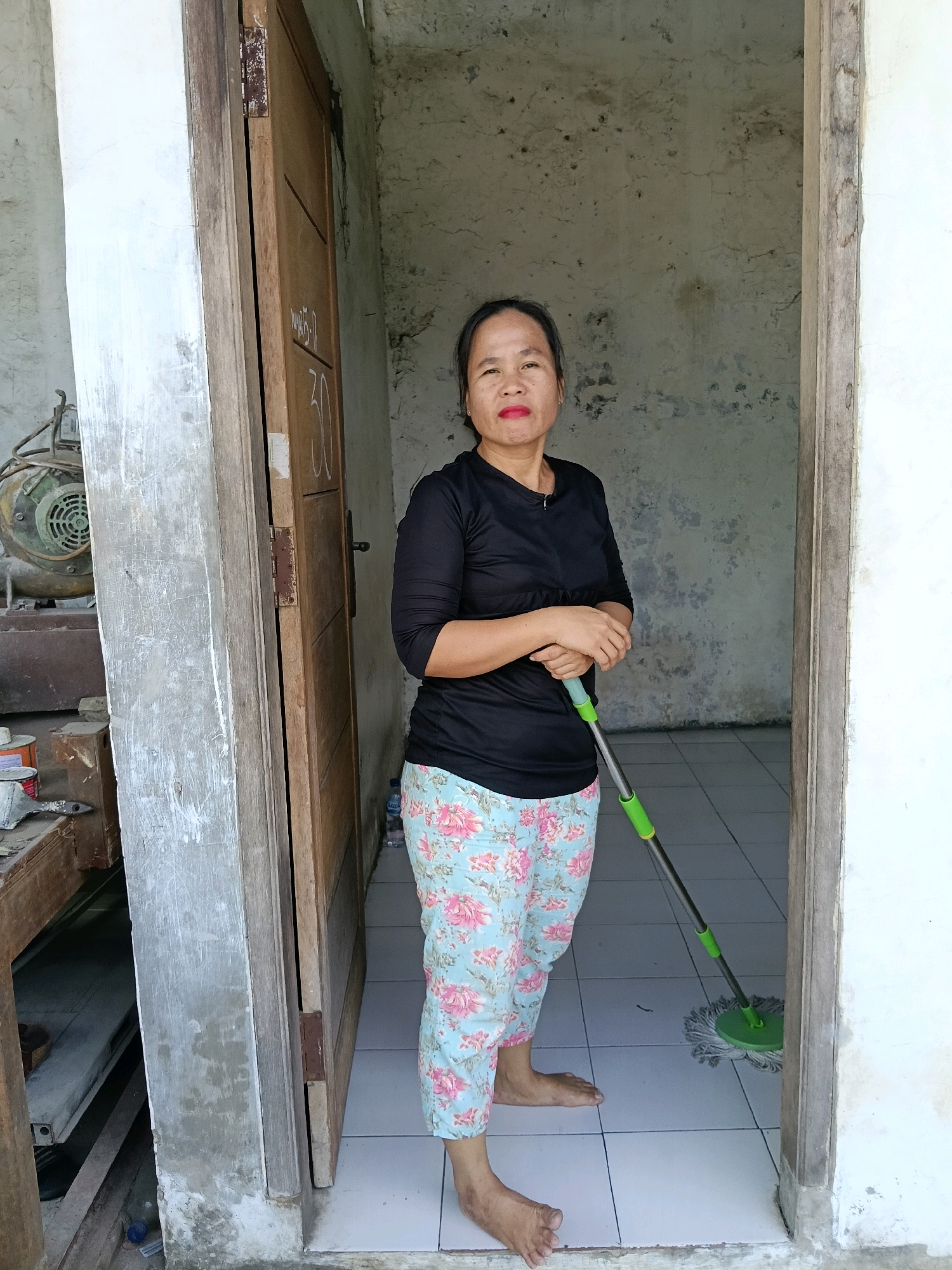 In the city of Belimbing, Indonesia, a woman stands in a doorway holding a mop. She is wearing a black long-sleeved shirt and floral-patterned pants. Her feet are bare. The doorway is made of wood, and a wooden door is partially open to her left, revealing a rough, textured wall behind it. To the left of the doorway, an industrial fan is visible. The floor is tiled in a light color. The overall scene appears to be a domestic setting, possibly a home or a workshop, given the presence of the fan and what looks like tools and cans on a workbench to the left. The wall behind the woman is unpainted and shows signs of age and wear.