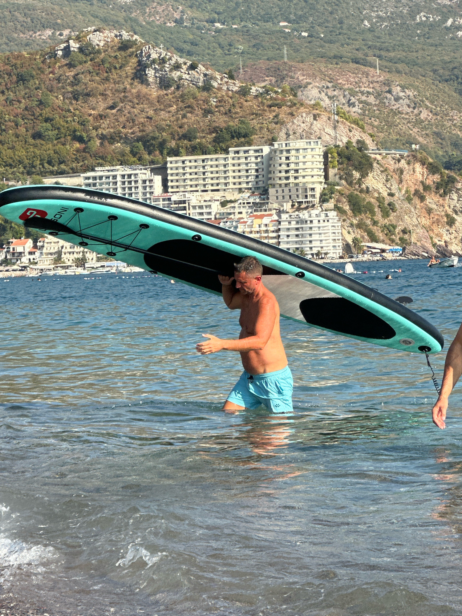 In the city of Boreti, Montenegro, a man is walking through shallow, clear water carrying a teal and black inflatable stand-up paddleboard on his shoulders. The man is wearing blue swim trunks. Another person's arm and hand are visible to the right of the frame, also in the water, with their hand extended. In the background, the blue sea is visible, dotted with several small boats and a white inflatable structure. Beyond the water, a densely populated coastline features multi-story buildings and a hillside covered in trees and rocky outcrops. A cross is visible on the highest rocky peak. The sun is shining, casting a bright light on the scene.
