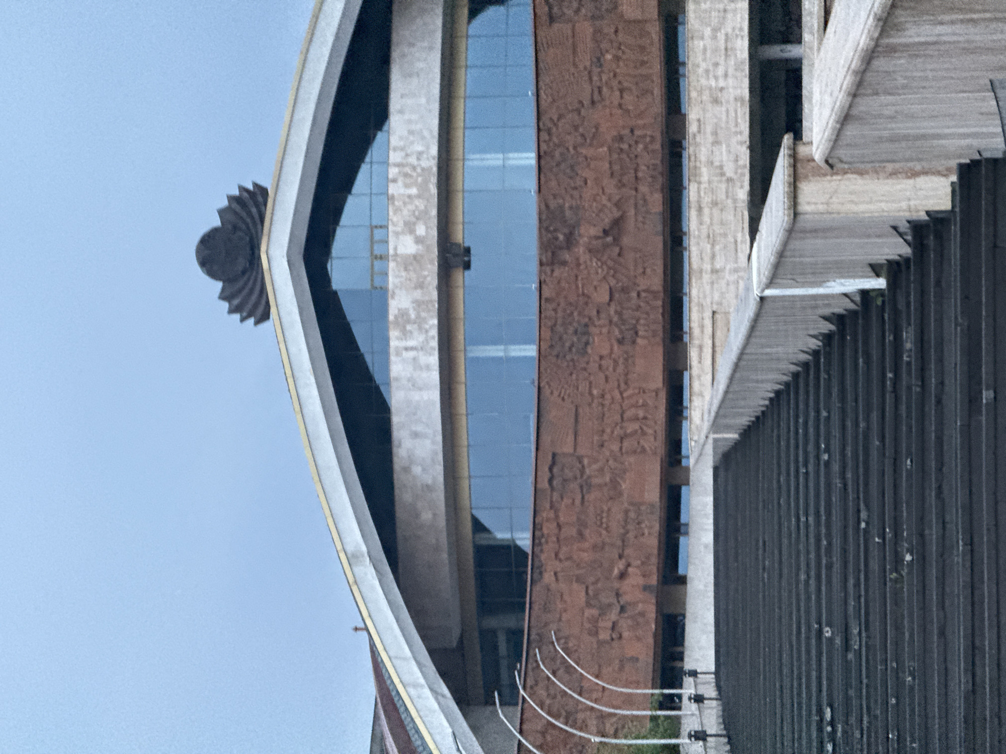 The image shows a partial view of the Karen Demirchyan Sports and Concerts Complex in Yerevan, Armenia. The architecture features a distinct design with a large, curved structure, a decorative flower-like element at the top, and multiple building materials including brown stone, glass, and grey textured paneling. A series of white power cables are visible. The sky in the background is light blue.