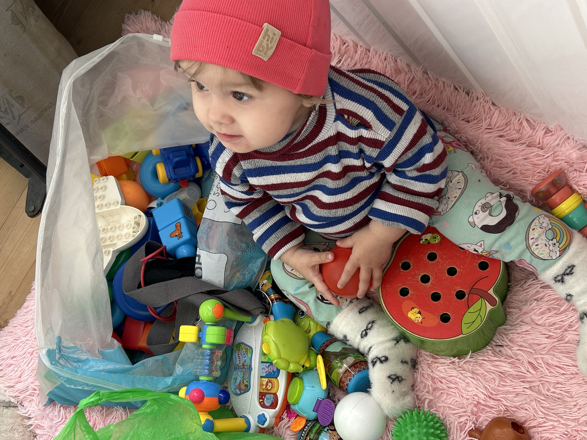 A baby or toddler sits on a fluffy pink rug, surrounded by a large collection of colorful toys. The child wears a coral-red knitted beanie with a "hi by" label, a striped long-sleeve top in blue, red, and white/grey, and light blue patterned pants featuring donuts and other motifs. The child is holding a red ball and looking with a curious and engaged expression slightly upwards and to the right.
Most of the toys are contained within a large clear plastic bag, with some spilling out onto the rug. Visible toys include various building blocks (blue, orange, white), a blue toy truck, a light green frog-shaped toy, a white toy piano with animal buttons and the words "HAPPY DAY" partially visible, a wooden watermelon-shaped lacing toy with black holes, a green spiky ball, a white ball, and a brown plastic toy. A green plastic bag is also visible on the bottom left.
The setting appears to be indoors, likely a home, with a light-colored background that could be a curtain or wall, and a wooden floor visible in the upper left. In the background, on a textured object, partial Cyrillic text "ФАБРИКА" (FABRIKA) can be seen. The overall scene suggests a playful and busy environment of a young child.