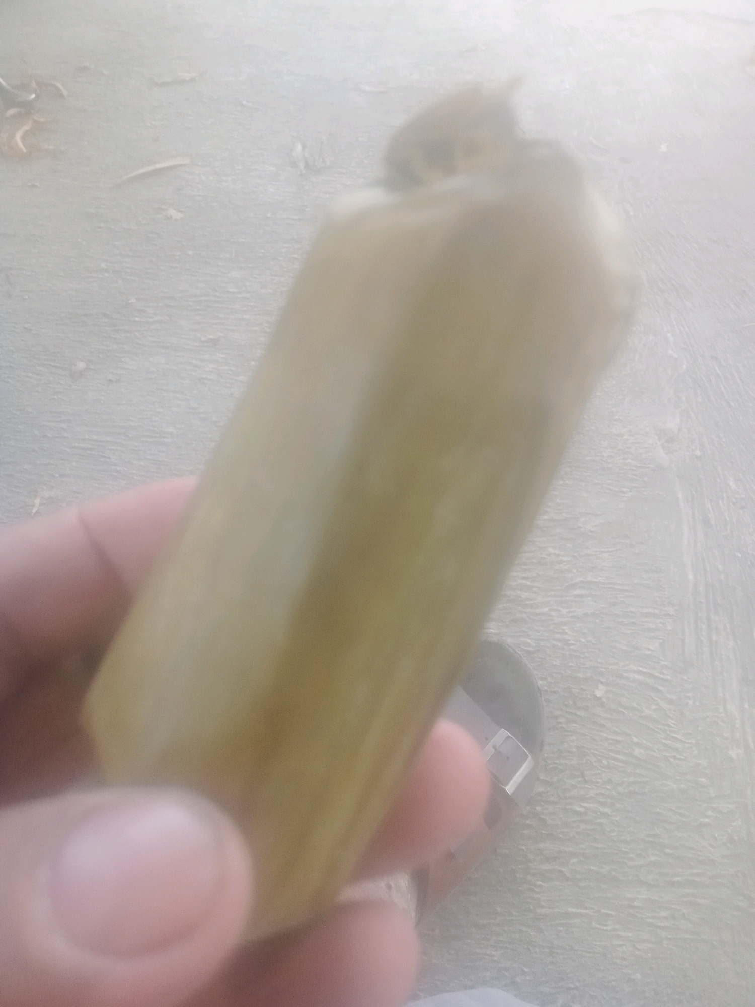 In Lebaksiu, Indonesia, a hand is holding a partially blurred, yellow, cylindrical object. The object appears to be some type of food item, possibly a piece of candy or a preserved fruit. The background consists of a grey, textured surface, possibly concrete or asphalt, with some dry leaves scattered on it. A portion of a dark shoe is visible in the lower right corner of the image. The lighting is bright and slightly overexposed, making it difficult to discern finer details.