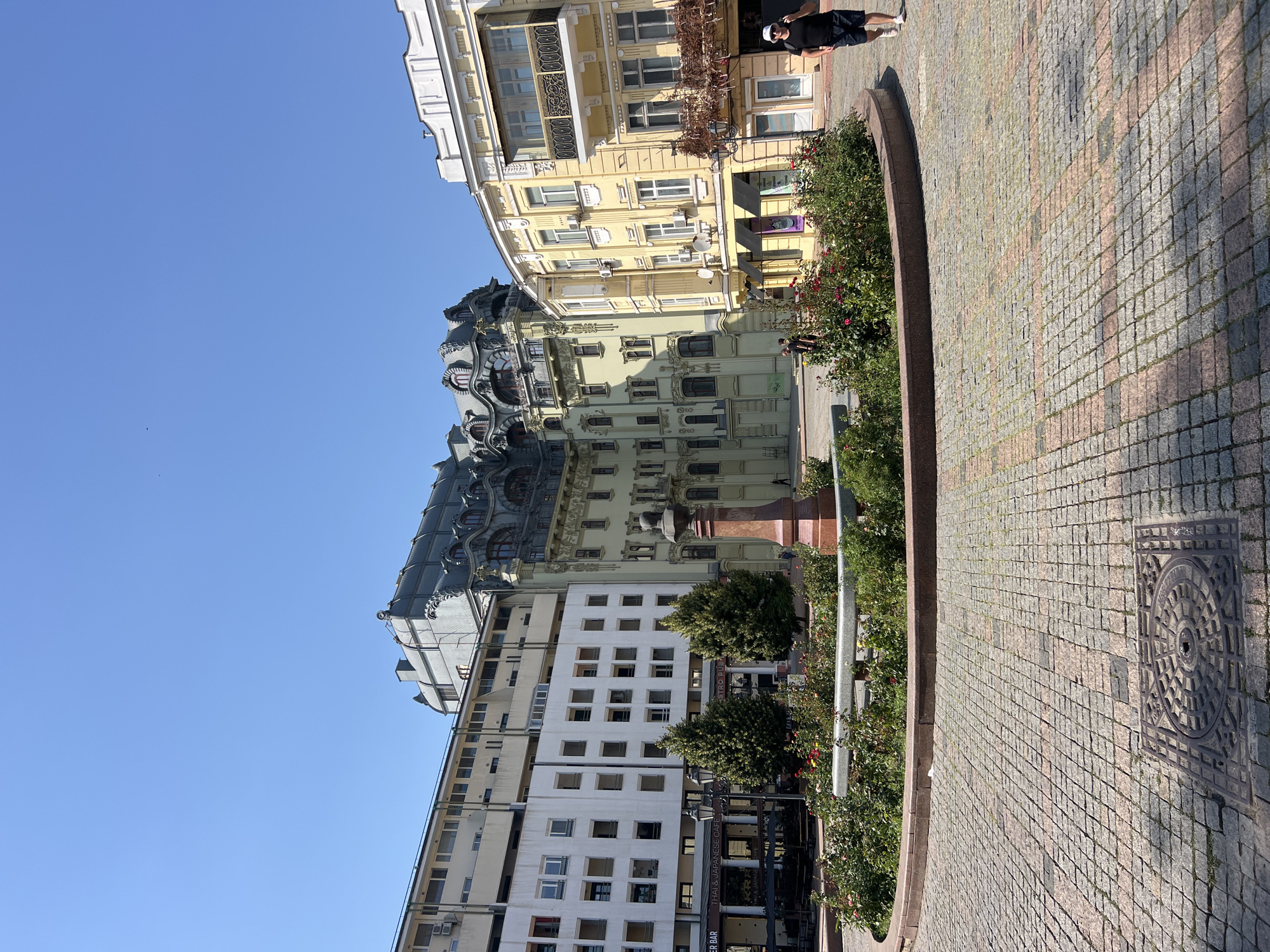 The image depicts a sunny day in an urban square in Odesa, Ukraine. In the foreground, a circular flower bed filled with roses and other greenery is bordered by a low, dark stone wall. A small, reddish fountain structure stands within the flower bed. The ground is paved with cobblestones in a diamond pattern, with a metal grate set into the pavement.
In the mid-ground, several buildings of varying architectural styles are visible. To the left, a modern, white building with many windows dominates the background. Adjacent to it is a building with a distinctive, ornate dark gray roofline and pale green facade. To the right, a bright yellow building with balconies and decorative elements stands.
Several trees are planted near the flower bed, and lampposts are present. In the background, people are walking. One man, wearing a black t-shirt, dark shorts, and a white baseball cap, is walking away from the camera towards the right side of the frame. The sky is clear and blue.