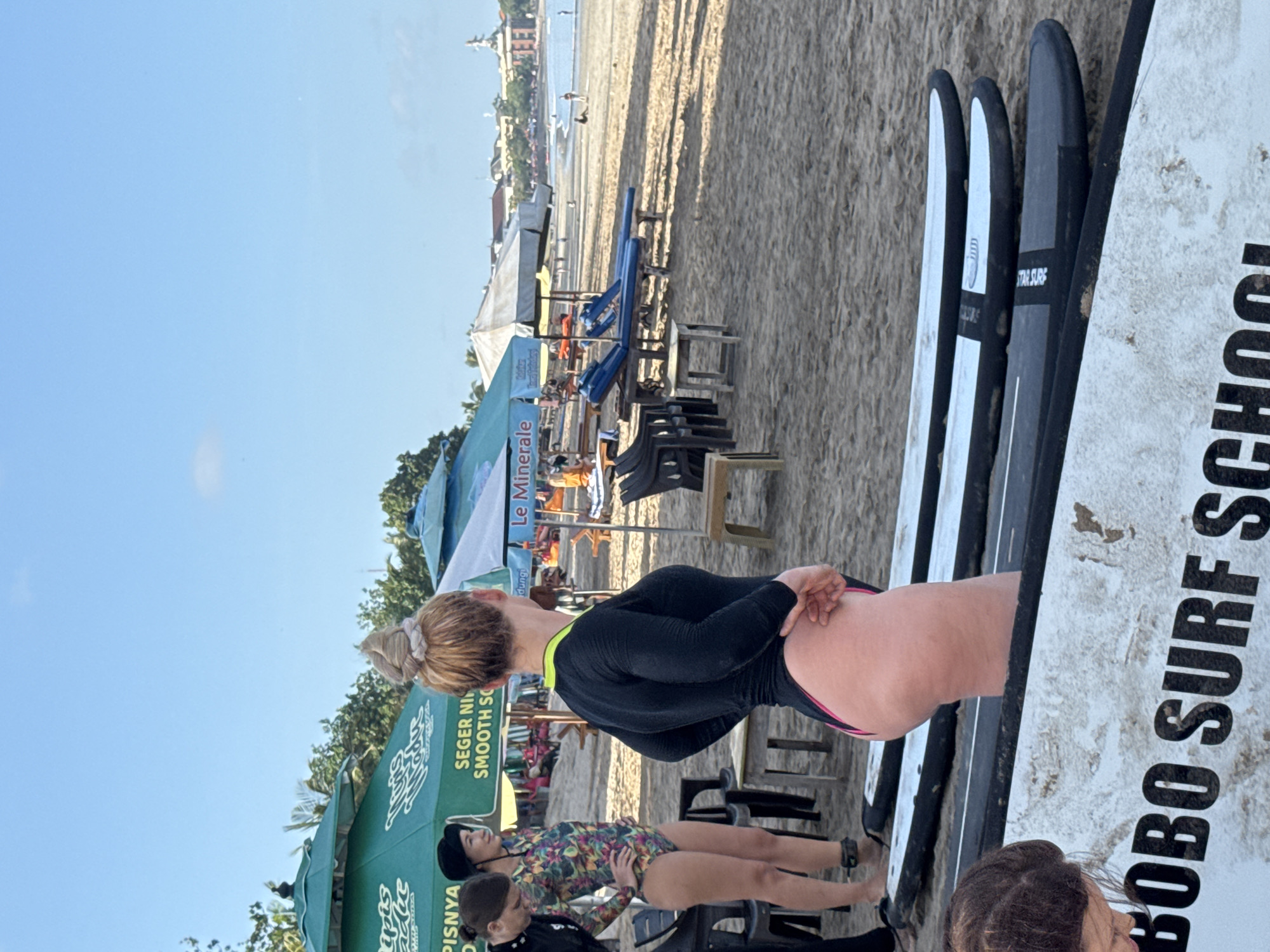 The image captures a scene at Kuta Beach in Kuta, Bali, Indonesia. The beach is sandy and visible in the background, with buildings and foliage lining the distant shore. Several individuals are present in the foreground, including a woman with blonde hair in a bun, wearing a black and neon yellow long-sleeved one-piece swimsuit. She is standing next to surfboards, one of which reads "BOBO SURF SCHOOL" in large black letters. Another individual is wearing a dark-colored swim suit that has multi-colored flowers on it. Green tents or canopies with the writing "SEGER NI" and "SMOOTH SO" are visible. There are also multiple chairs lined up near the tents. Several other people are partially visible, adding to the scene's complexity. The sky is clear and blue, suggesting a sunny day.