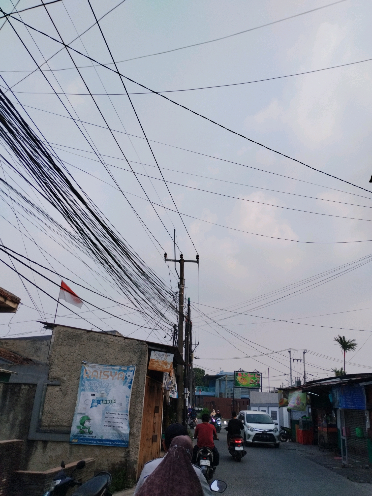 This image depicts a street scene in Bojong Gede, Indonesia, characterized by a dense network of overhead power lines crisscrossing the sky. The time appears to be late afternoon or early evening, with an overcast sky. In the foreground, a motorcycle is parked on the left. A building with a textured, light-colored facade is prominent on the left, featuring a poster for "RAISYA Laundry" with pricing information and contact details. A small Indonesian flag is visible, mounted on a pole near the building.
The street is paved and appears to be a two-lane road. Several motorcycles are in motion, with riders heading away from the viewer. A white car is also traveling down the street. On the right side of the road, there are small shops or stalls with open fronts, displaying various goods. A palm tree is visible in the background to the right. The overall impression is a typical urban or semi-urban street in Indonesia, with a focus on daily life and commerce.