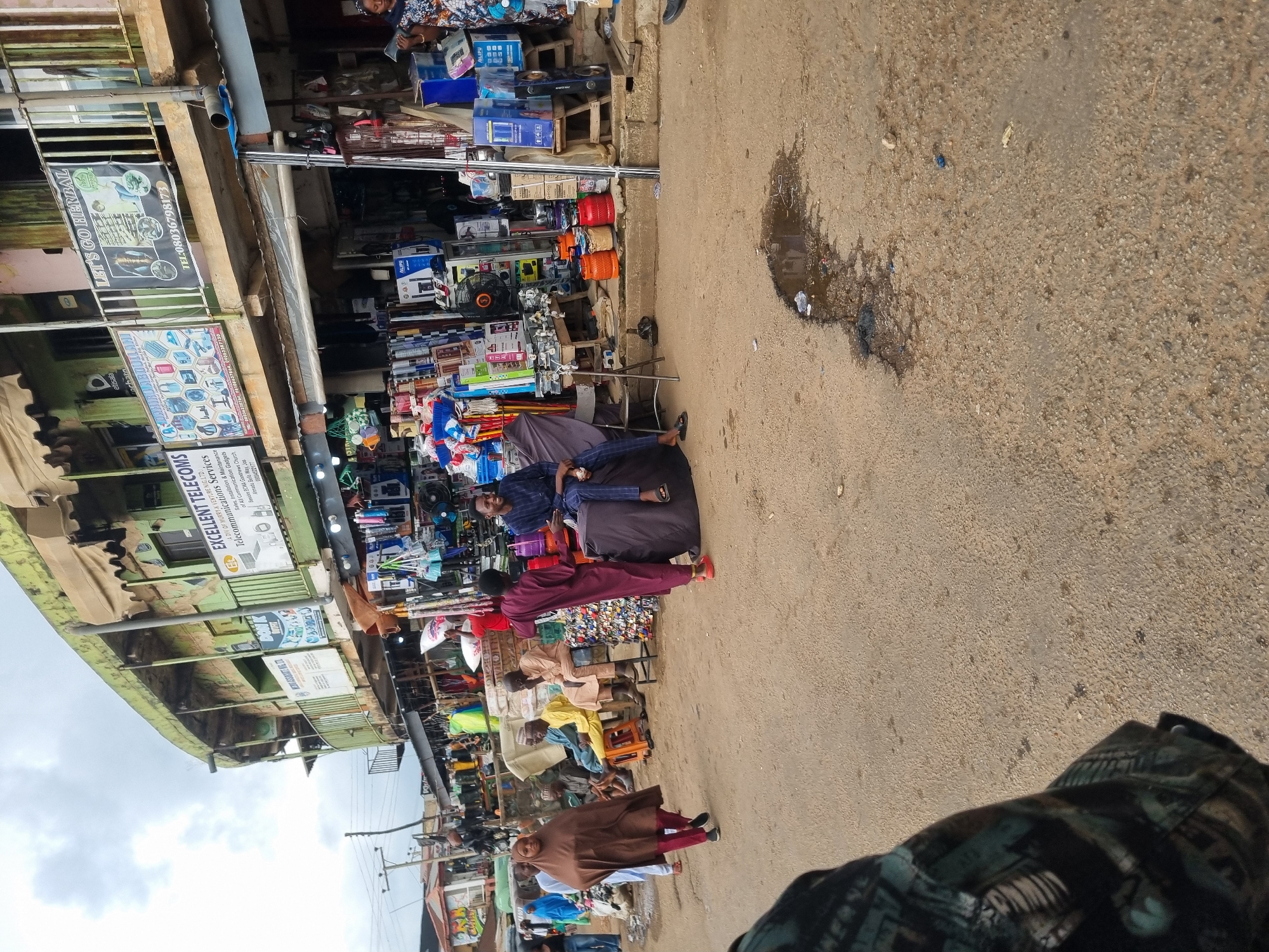 The image depicts a street scene in Jos, Nigeria, likely a market area. In the foreground, a person wearing a patterned shirt is partially visible on the left, with their arm and shoulder taking up the bottom left corner. The ground is a dusty, worn asphalt surface with a dark, wet patch with debris in the mid-ground.
In the background, a row of shops and stalls are set up along a multi-story building. The building has a weathered, green facade and multiple balconies. Various goods are displayed for sale, including electronics, household items, and what appear to be gas cylinders stacked in orange and red. Several people are present in the scene.
On the left, a woman in a brown hijab and matching trousers walks away from the camera. To her left, another woman in a blue hijab stands. Several men are seated or standing near the shops. One man in a yellow outfit sits on an orange stool, while another in a light brown robe is standing. Further to the right, a man in a dark maroon robe stands near a large, dark-covered object, possibly a seating area or display. He is interacting with a man in a blue and white striped outfit, who is seated on the dark object. Another man in a dark patterned shirt is visible further to the right, seated and smiling.
Signs in English are visible on the buildings, including "EXCELLENT TELECOMS" and "LET'S GO HERBAL," suggesting a mix of businesses operating in the area. The sky is overcast with a muted, grey tone. The overall atmosphere suggests a busy commercial hub.