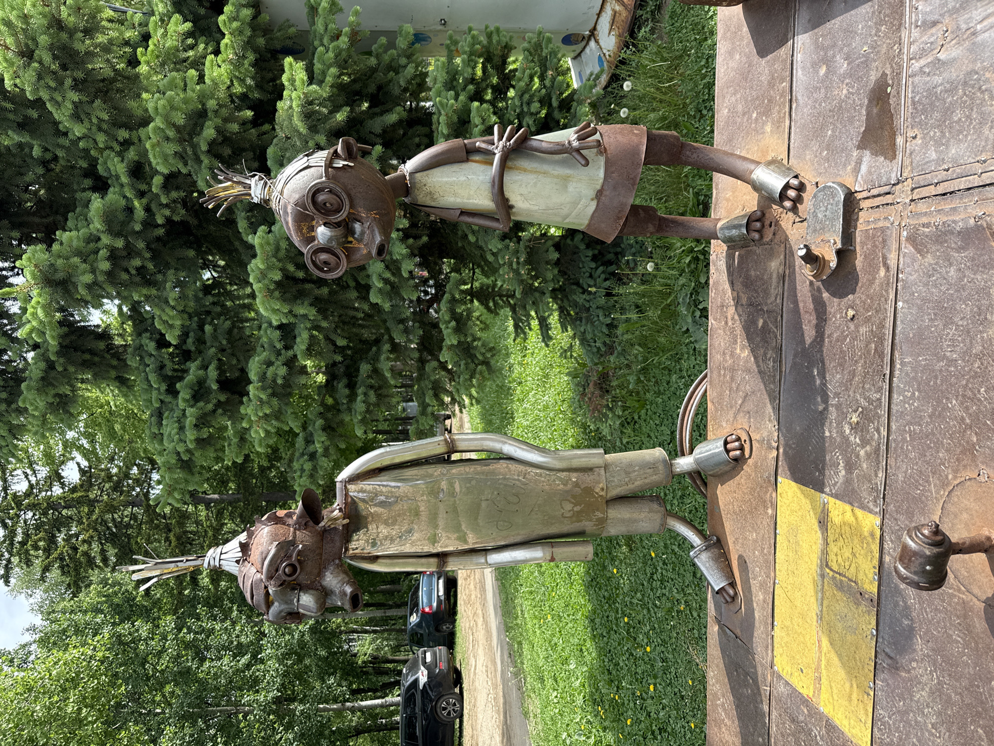 The image shows two metal sculptures of humanoid figures. The sculptures are constructed from various metal parts and are located outdoors. One sculpture is standing on a metal surface with a yellow square. The environment appears to be a park or green space. The location is likely Izhevsk, Russia, given the prompt.
