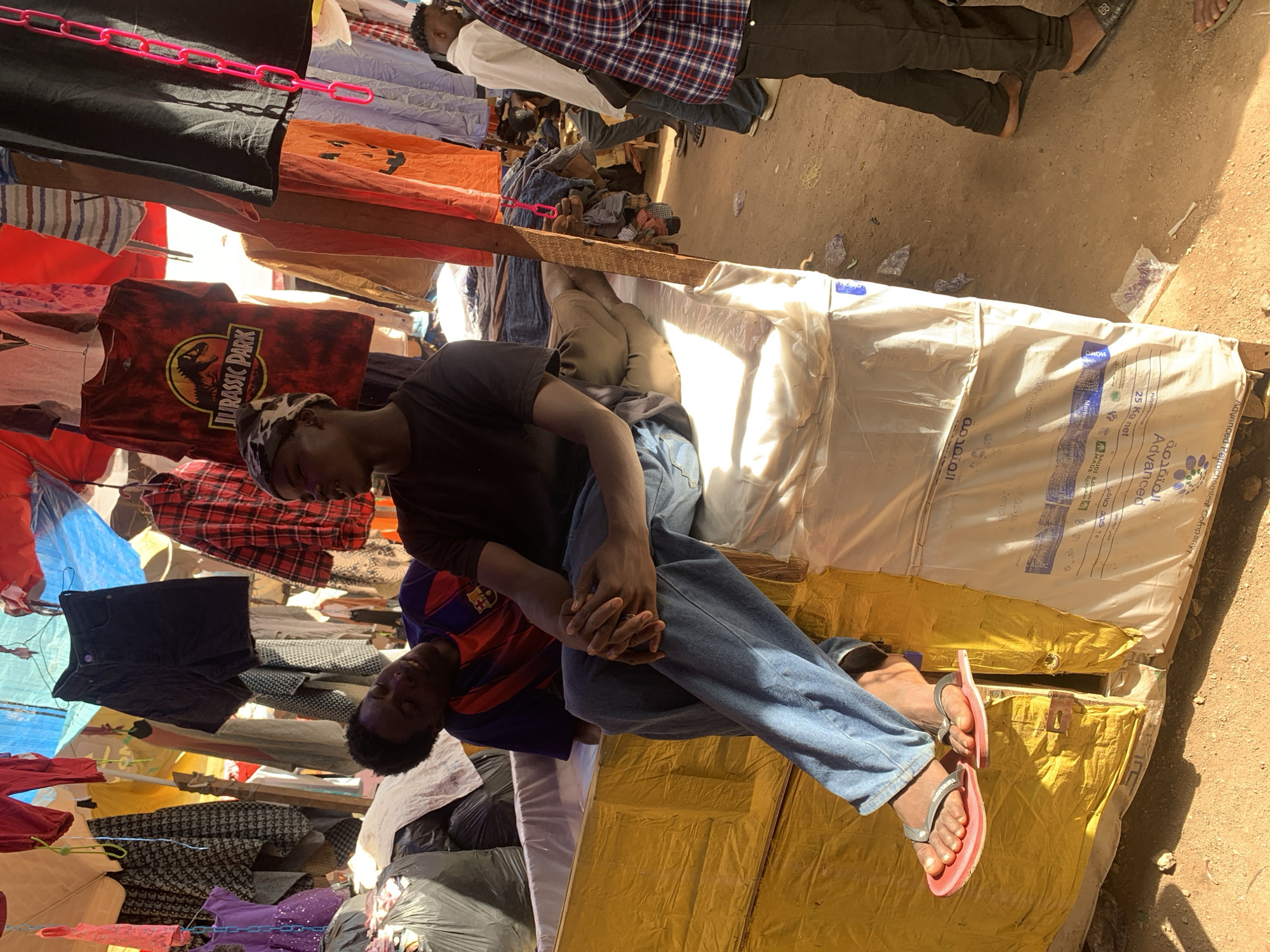 The image, likely taken in Jos, Nigeria, shows a street scene with vendors displaying clothing for sale. Clothes hang from makeshift racks, creating a dense, colorful display. A man wearing jeans and a black T-shirt, with a cap or head covering, sits on what appears to be large sacks or bags, possibly filled with goods. He is interacting with another man who is wearing a striped shirt. The ground is dirt or soil, suggesting an outdoor market setting. The image has a slightly tilted perspective. Several of the items of clothing have popular logos or brand names.