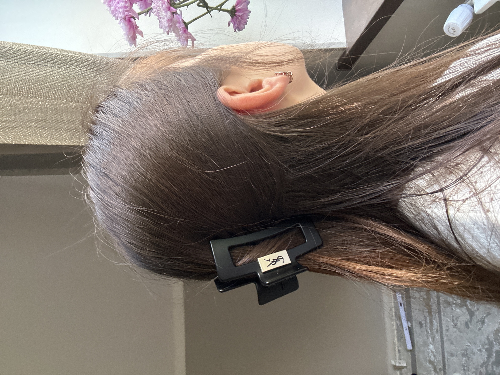 A close-up, side-profile shot captures the back of a person's head and ear, adorned with a black claw clip featuring a prominent gold logo. The person's brown hair is visible, styled with the clip securing a portion of it. The background includes a neutral-colored wall, a sheer curtain, and a glimpse of pink flowers. To the lower left, a textured surface is visible with a white object and a thin horizontal line. The overall lighting suggests an indoor setting near a window. The location is identified as a residential area in Кызыл-Булак, Kyrgyzstan.