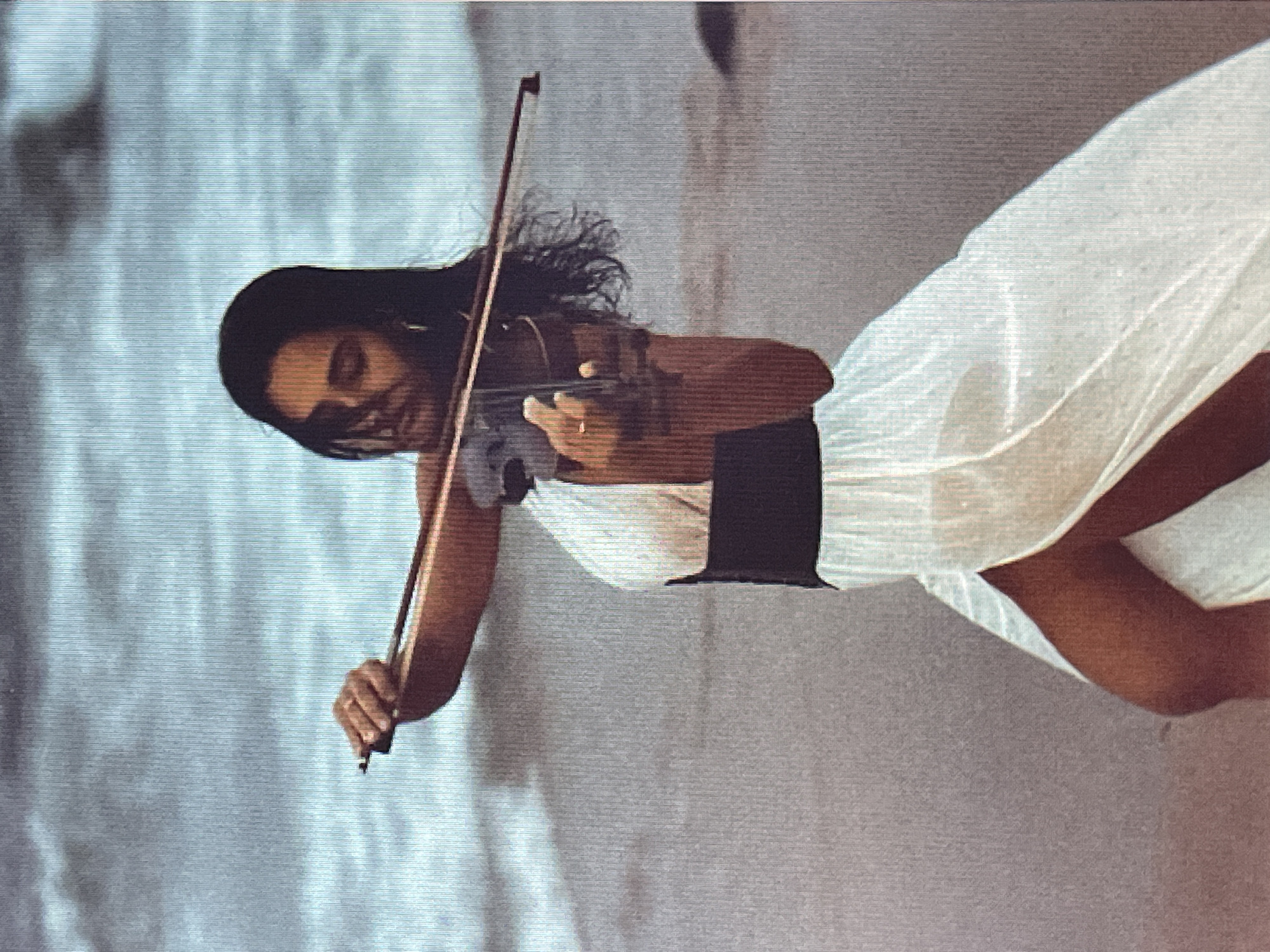 A woman with dark hair is playing the violin. She is wearing a white dress with a black belt and a black underskirt. The setting appears to be outdoors, possibly on a beach, with sand and water visible. The woman is looking down at the violin as she plays. Her expression is one of concentration. The lighting suggests it could be daytime. The overall mood of the image is serene and artistic.