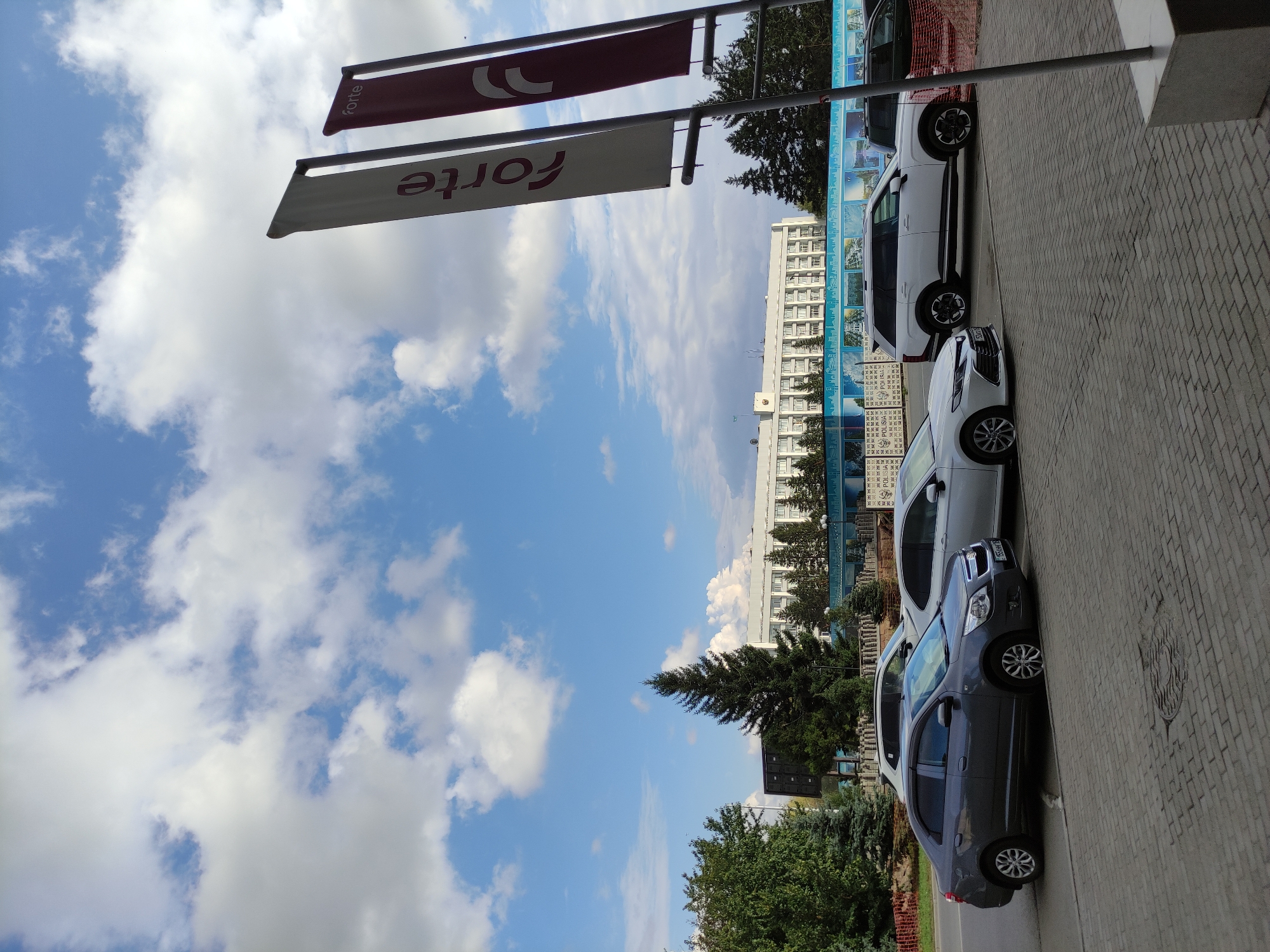 The image depicts an outdoor scene in Semey, Kazakhstan, on a partly cloudy day. In the foreground, a grey sedan and a white SUV are parked on a paved area adjacent to a brick sidewalk. Two large banners, one white and one burgundy, are visible in the right portion of the frame, bearing the word "Forte" in red lettering.
In the middle ground, several evergreen trees are situated in front of a large, white, multi-story building with numerous windows. A blue mural or banner with a cityscape design runs along the base of this building. Behind the trees and building, the sky is a mix of blue and white clouds. The scene appears to be a street or a parking area in front of a commercial or administrative building. No people are visible in the image.