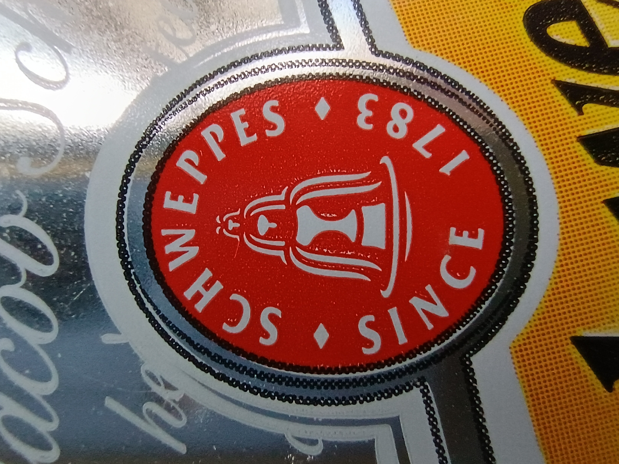 The image shows a close-up of a Schweppes logo, likely on a product or advertisement. The logo is circular and features the text "SCHWEPPES SINCE 1783" in white lettering against a red background. A stylized emblem, possibly representing a fountain or a historical figure, is visible in the center of the logo. The logo is surrounded by a black and silver beaded border. The background is a metallic silver with blurred white text and a yellow textured area at the bottom with black lettering partially visible. There is no indication of the city of Zaporizhzhia, Ukraine in the image.
