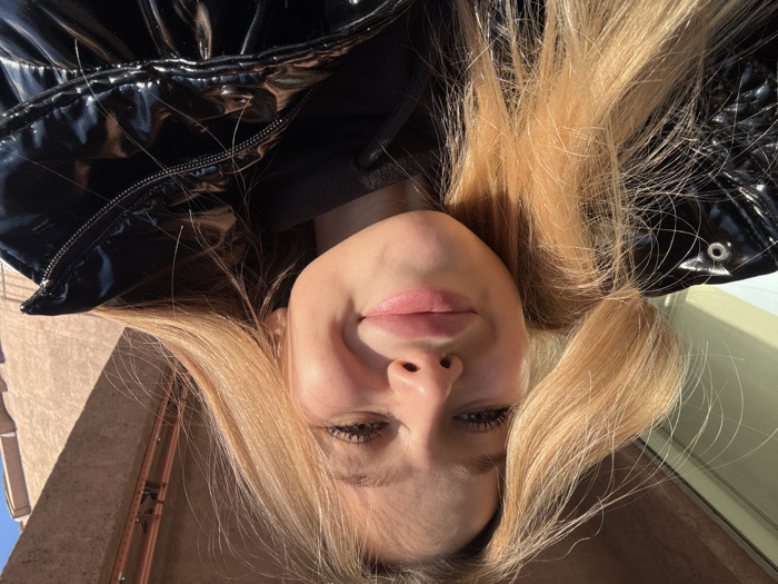 A close-up, low-angle outdoor photograph features a young female subject under bright sunlight. The individual has long, straight, light blonde hair, visible eyelashes, and full, pink-toned lips, displaying a closed-mouth smile. She is wearing a black, shiny, puffer-style jacket over a dark hooded sweatshirt. The background is out of focus: to the left, a bright, reflective surface is visible; to the right, a light brown building facade with a dark horizontal fixture is present, indicating an urban or suburban setting. The specific city location in Unknown, Unknown, is not discernible from the visual evidence.
